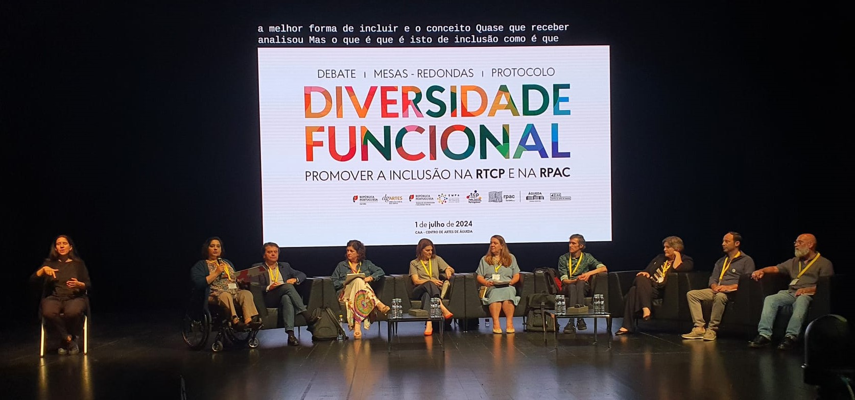 Palco com 10 pessoas sentadas de frente para uma plateia onde, entre elas, se encontra uma pessoa em cadeira de rodas e uma intérprete de Língua Gestual Portuguesa. Por detrás das pessoas vê-se uma tela grande onde está escrito, em cima, uma frase que legenda o que o orador estava a falar. Por baixo da frase encontra-se o texto “Debate | Mesas-Redondas | Protocolo” em letras pretas, assim como “DIVERSIDADE FUNCIONAL” e “PROMOVER A INCLUSÃO NA RTPC E NA RPAC”. O rodapé inclui alguns logótipos como o da República Portuguesa, da DGARTES, da EMPA, da RTPC e da RPAC. Abaixo dos logos, está o texto, 1 de julho de 2024, CAA – Centro de Artes de Águeda