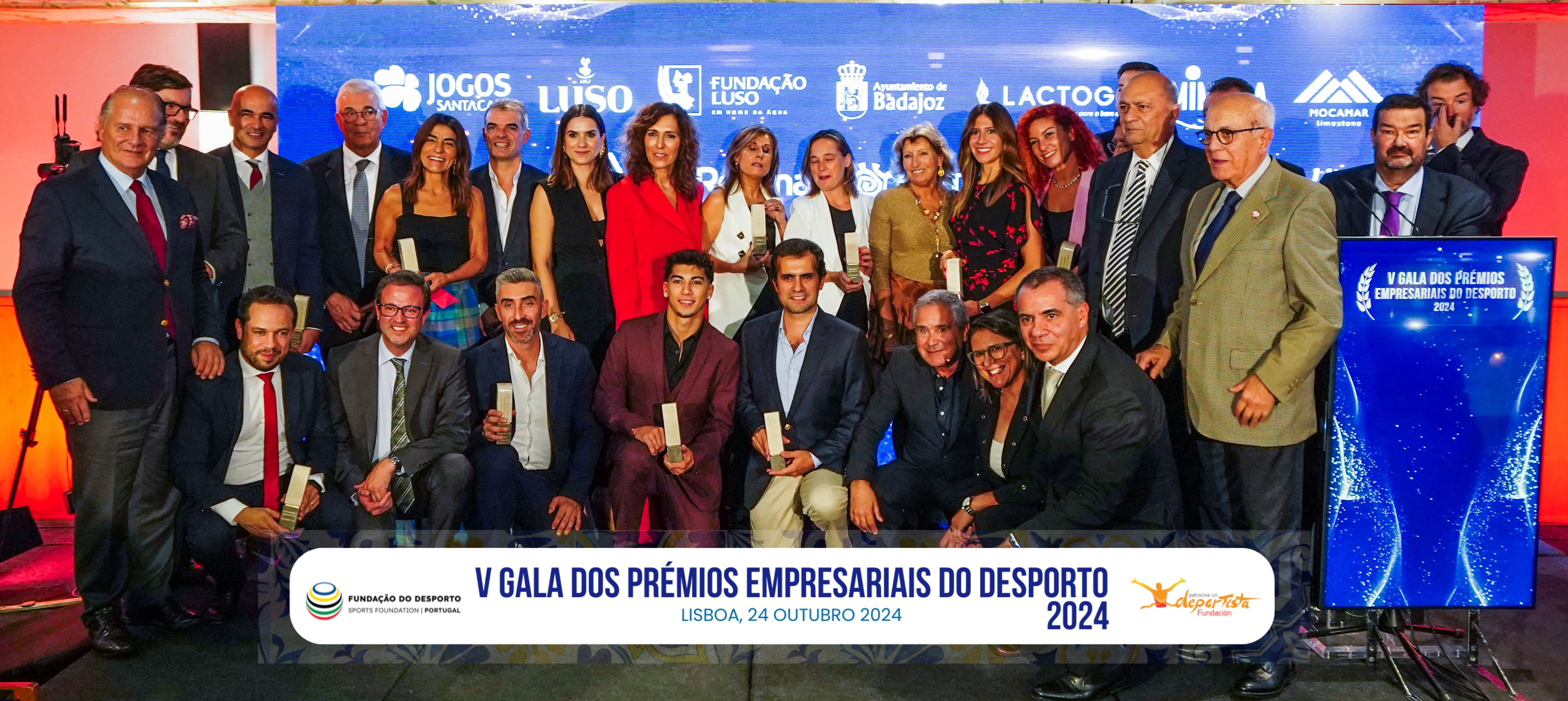 Foto de grupo com 27 pessoas, entre elas premiados e outros convidados que estiveram presentes no evento. Do lado direito é visível um púlpito azul com o texto “V edição da Gala dos Prémios Empresariais do Desporto, 2024”. Por detrás do grupo está um painel azul com logos de várias marcas.