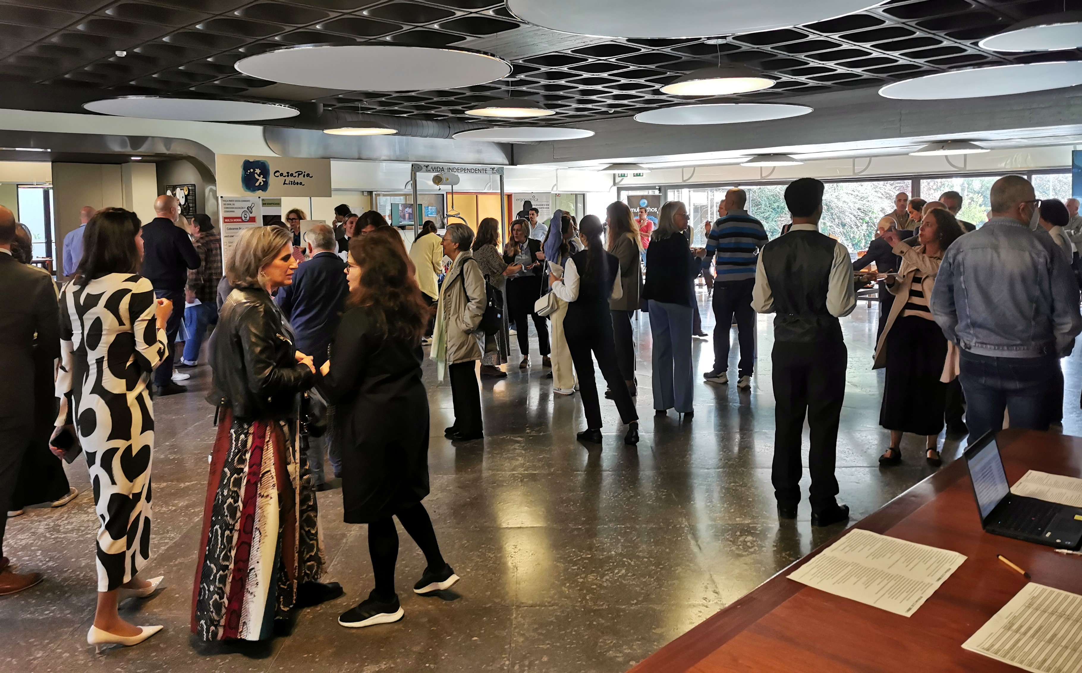 You see a group of people standing, interacting, in an atrium of a building. In the lower right corner, a table, a laptop and some sheets are visible.