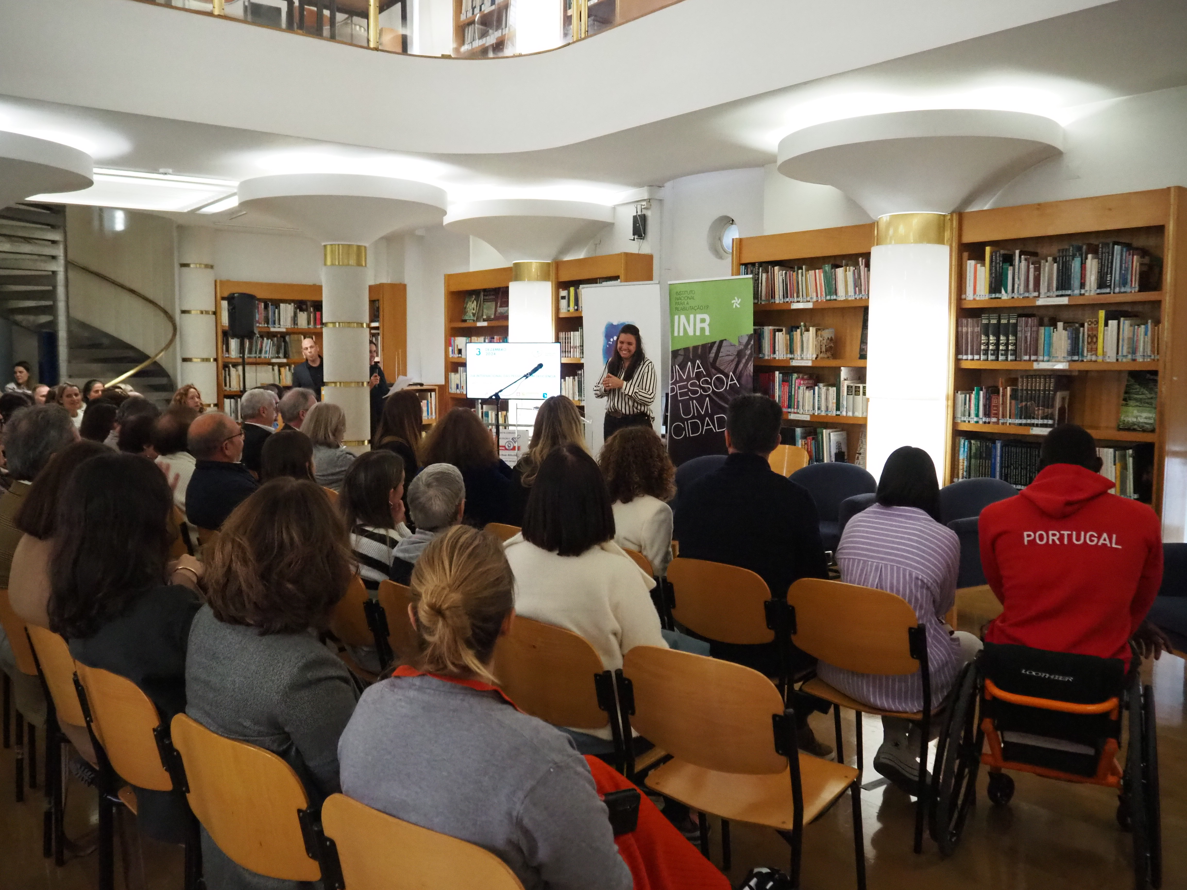 Público com várias pessoas sentadas a assistirem ao evento. A apresentadora é uma mulher e está a sorrir