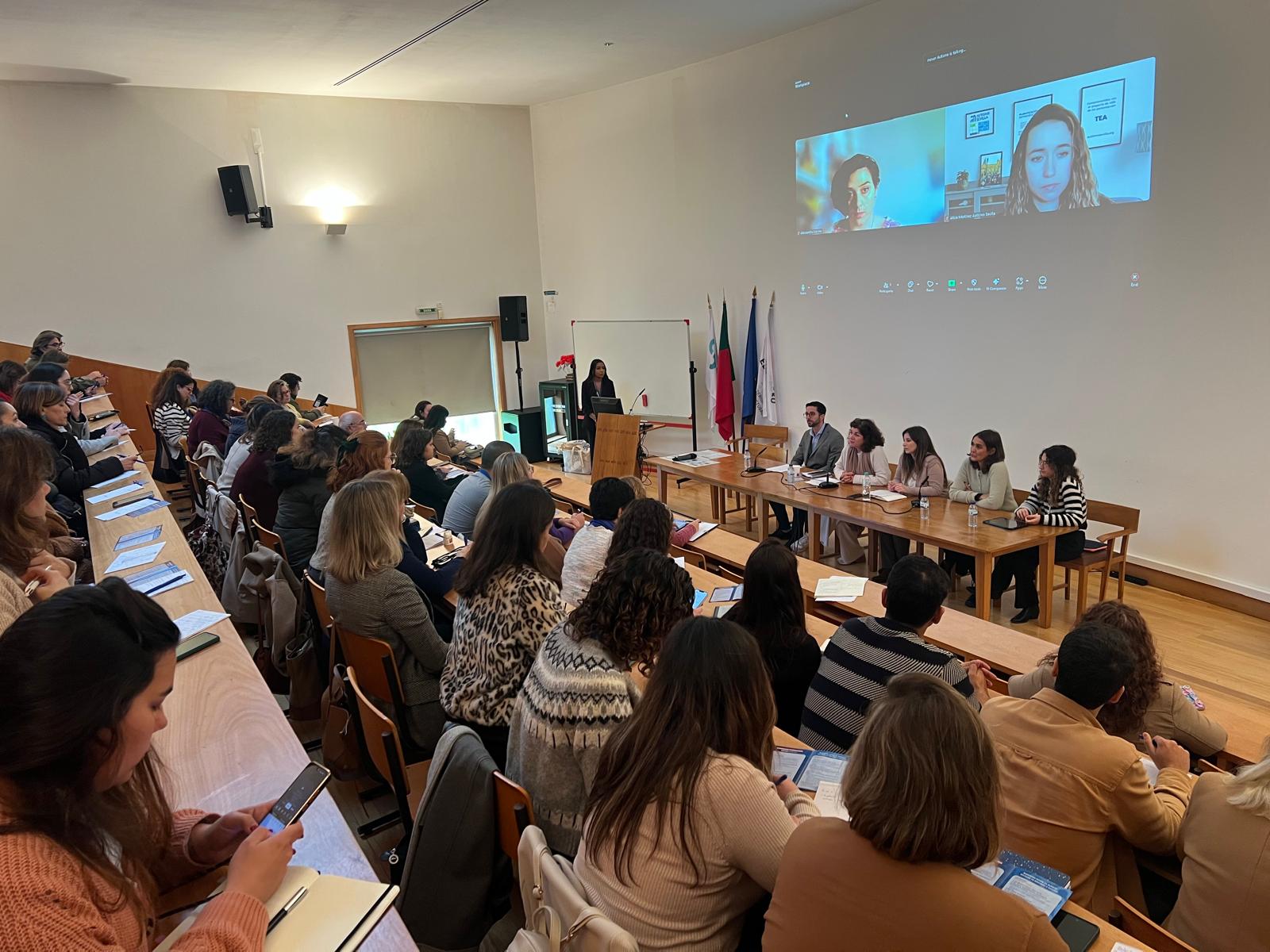 Há uma tela de projeção onde se vê duas pessoas em videochamada. Na frente da sala, há uma mesa com várias pessoas sentadas, entre elas Helena Alexandre e participantes da mesa-redonda sobre o projeto europeu “Self-Determination & Inclusion by Theather” (Autodeterminação e Inclusão pelo Teatro). É visível o auditório com várias pessoas presentes.