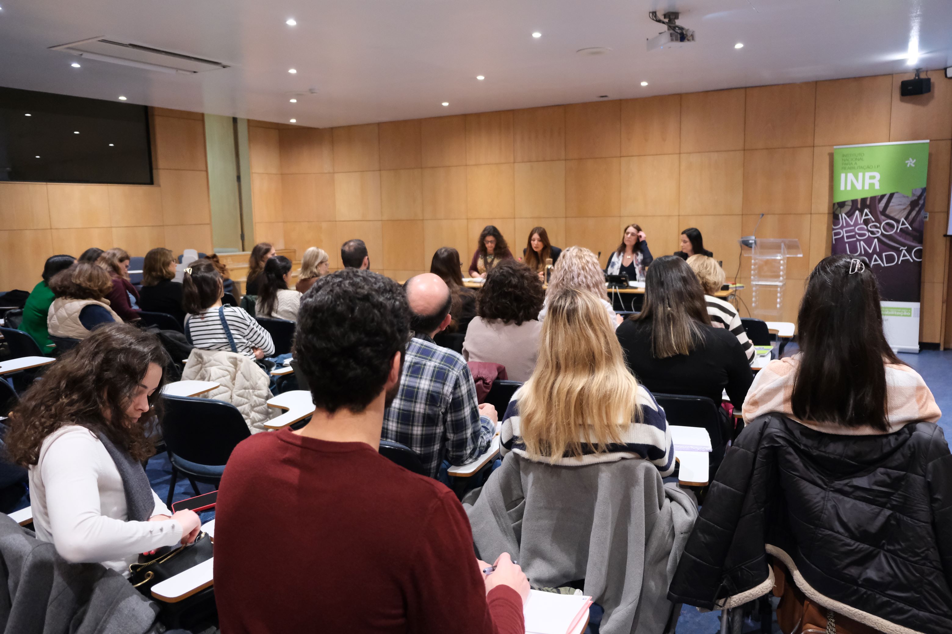  É visível um auditório com público a olhar para as quatro oradoras. Do lado direito delas, está um rollup do INR. 