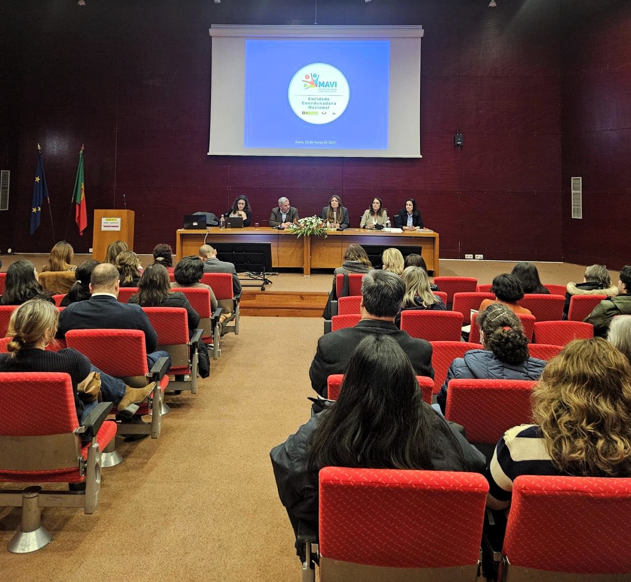  É visível um auditório com público, que está a olhar para 5 pessoas que estão sentadas à mesa. Sónia Esperto está a falar para audiência. Por detrás delas está uma projeção, que tem uma imagem que diz “MAVI, Entidade Coordenadora Nacional”. 