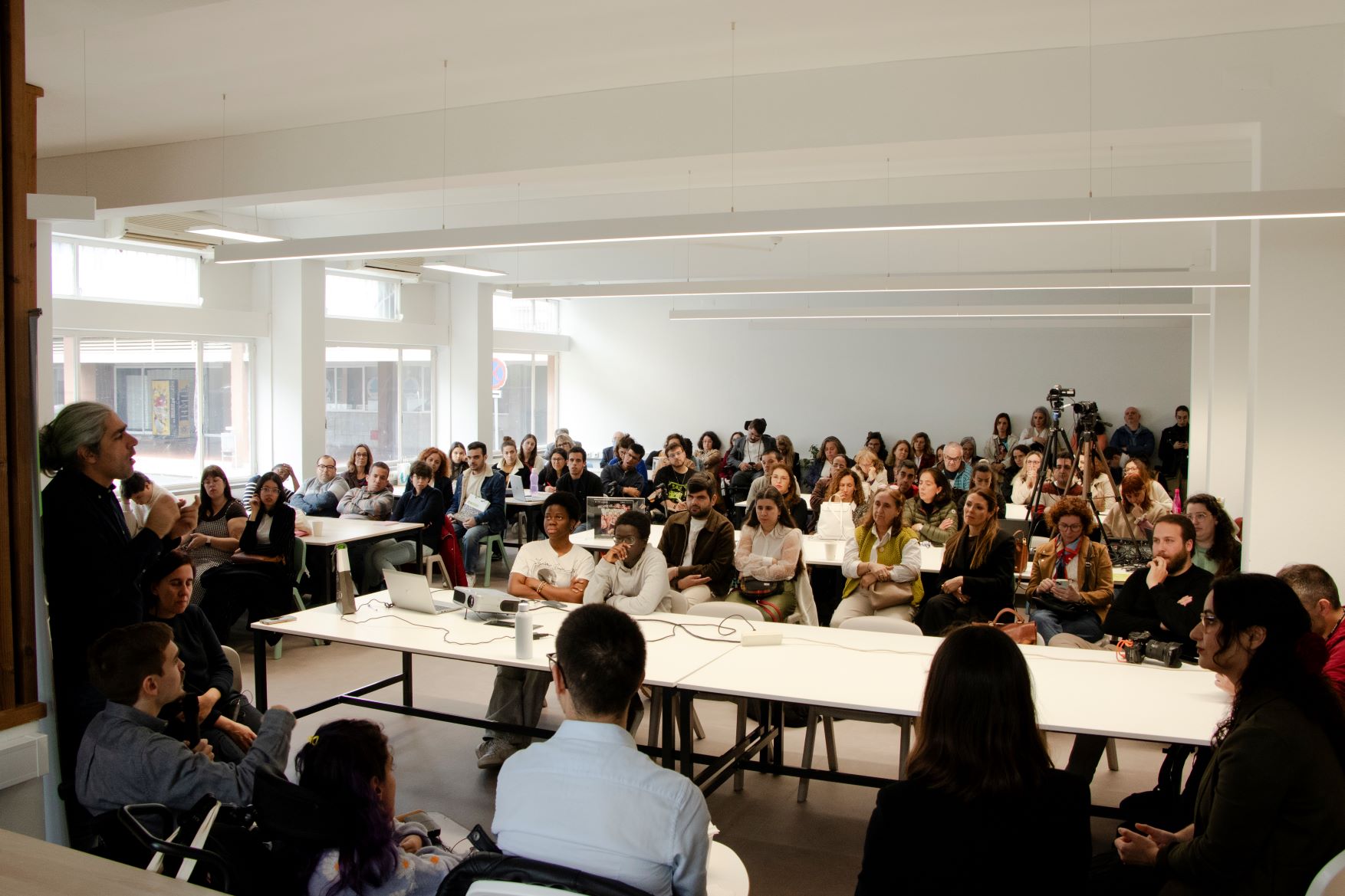  Várias pessoas sentadas, pertencentes ao público que está a assistir a uma mesa-redonda constituída por 5 participantes. Vê-se, também, dois intérpretes de Língua Gestual Portuguesa. 