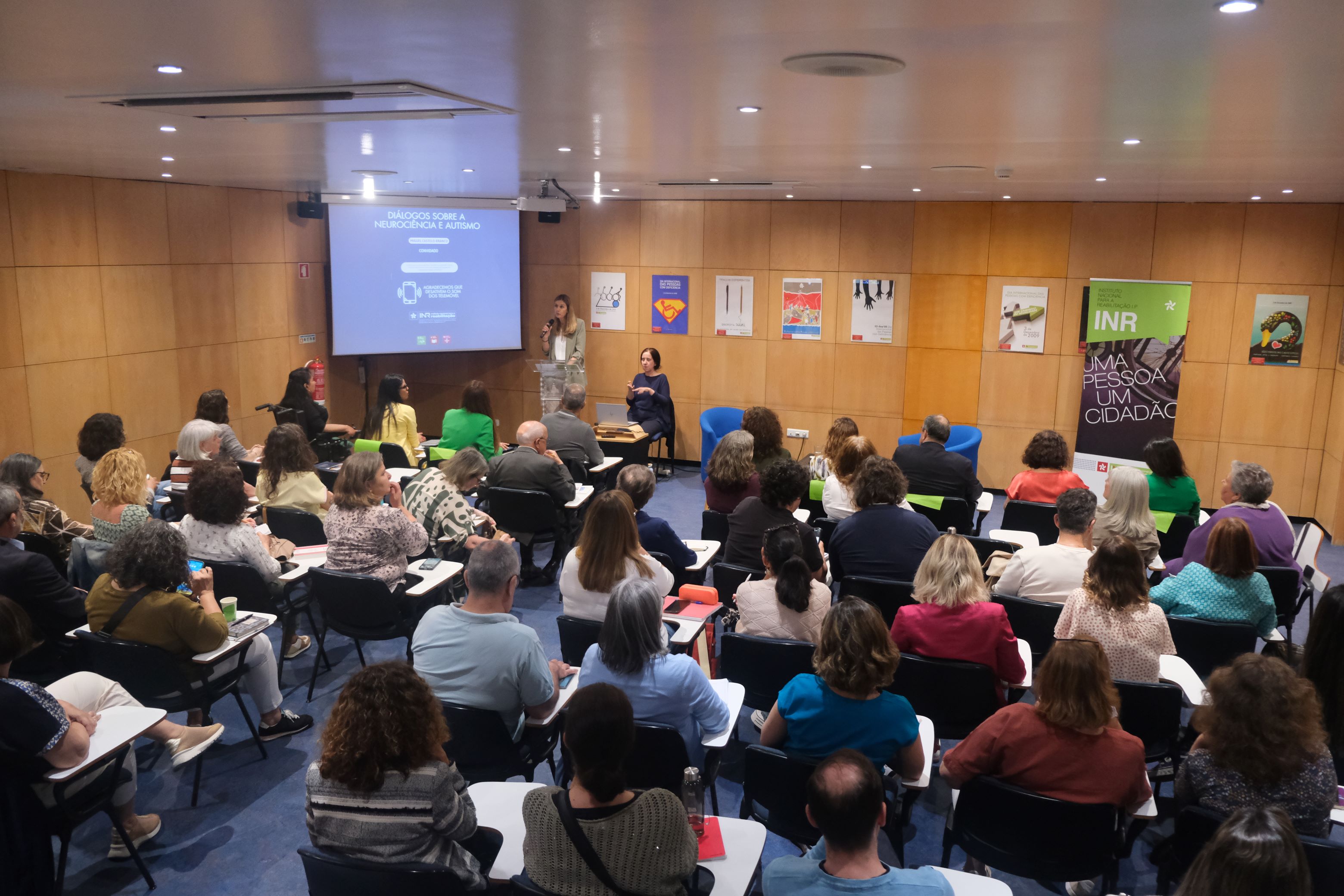 You can see an auditorium with an audience that completely fills the room. In the background of the image is visible a screen with projection, a person speaking in the pulpit and, next to her, a Portuguese Sign Language interpreter.