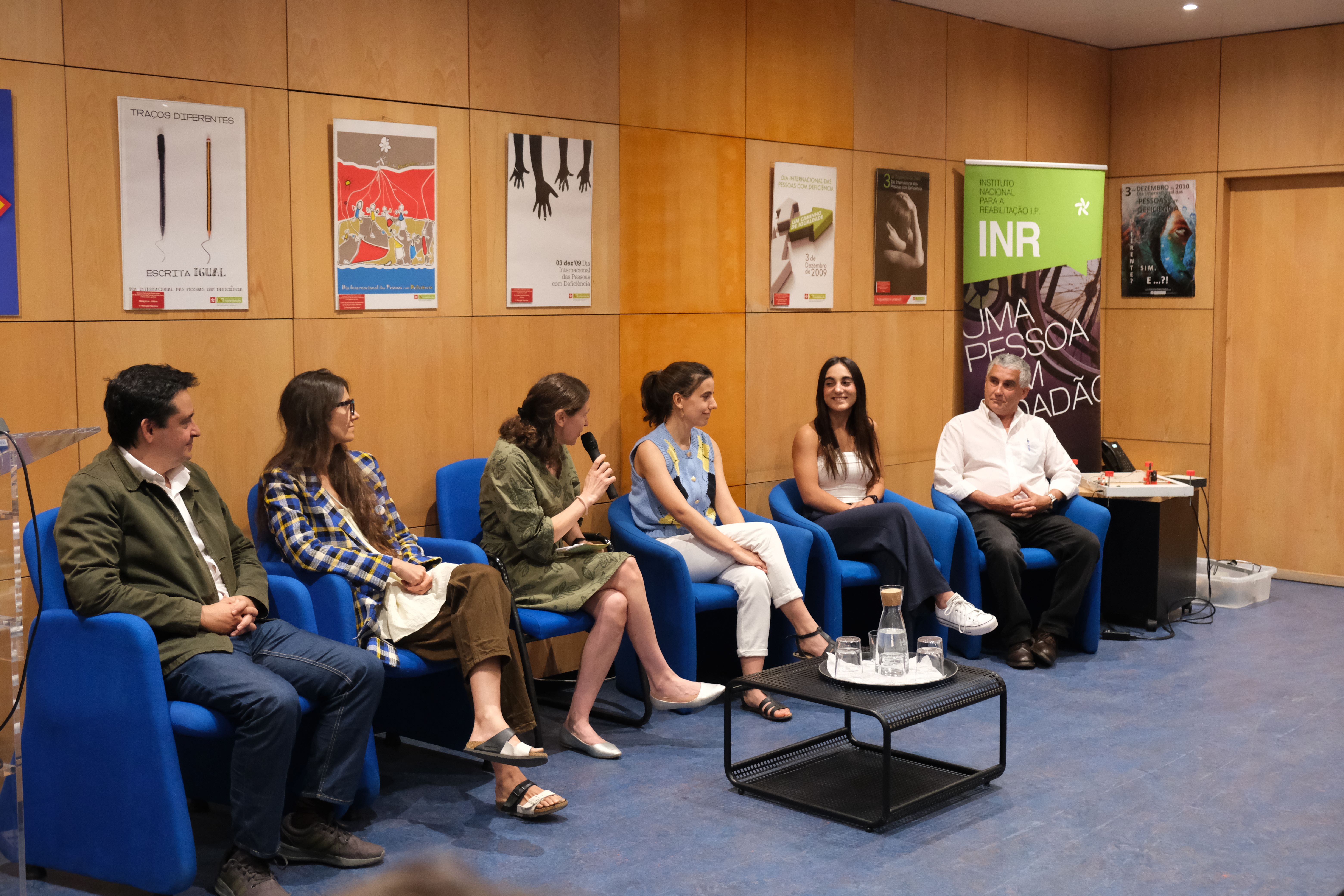 There are 6 people sitting, participants in the session, of the winning projects. One of the participants, the moderator, has a microphone in her hand talking.
