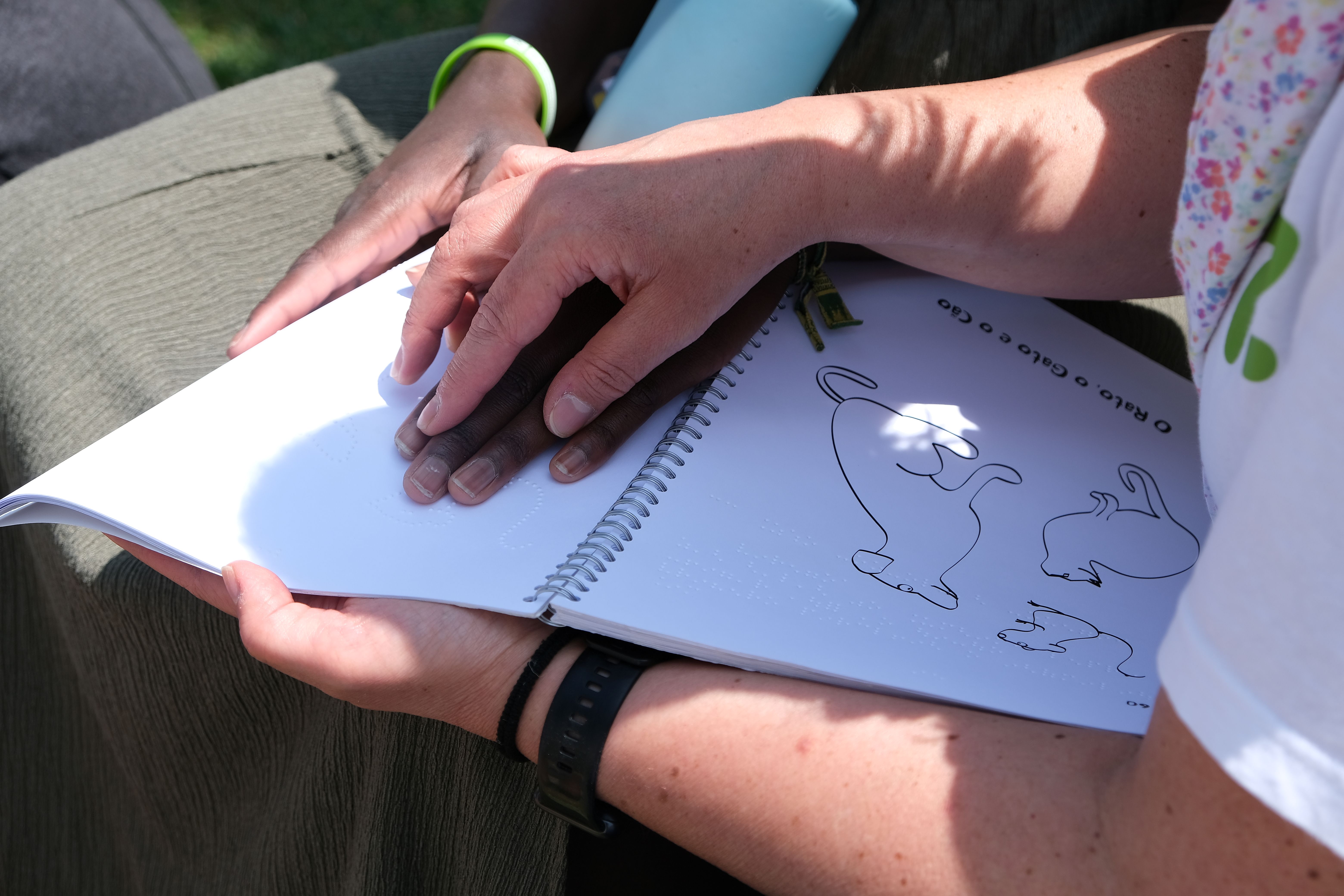  Foto onde se vê as mãos de uma pessoa a guiar as mãos de outra pessoa, surdocega, a ler um livro em braile. Photo where you see the hands of one person guiding the hands of another person, deafblind, reading a book in braille. 