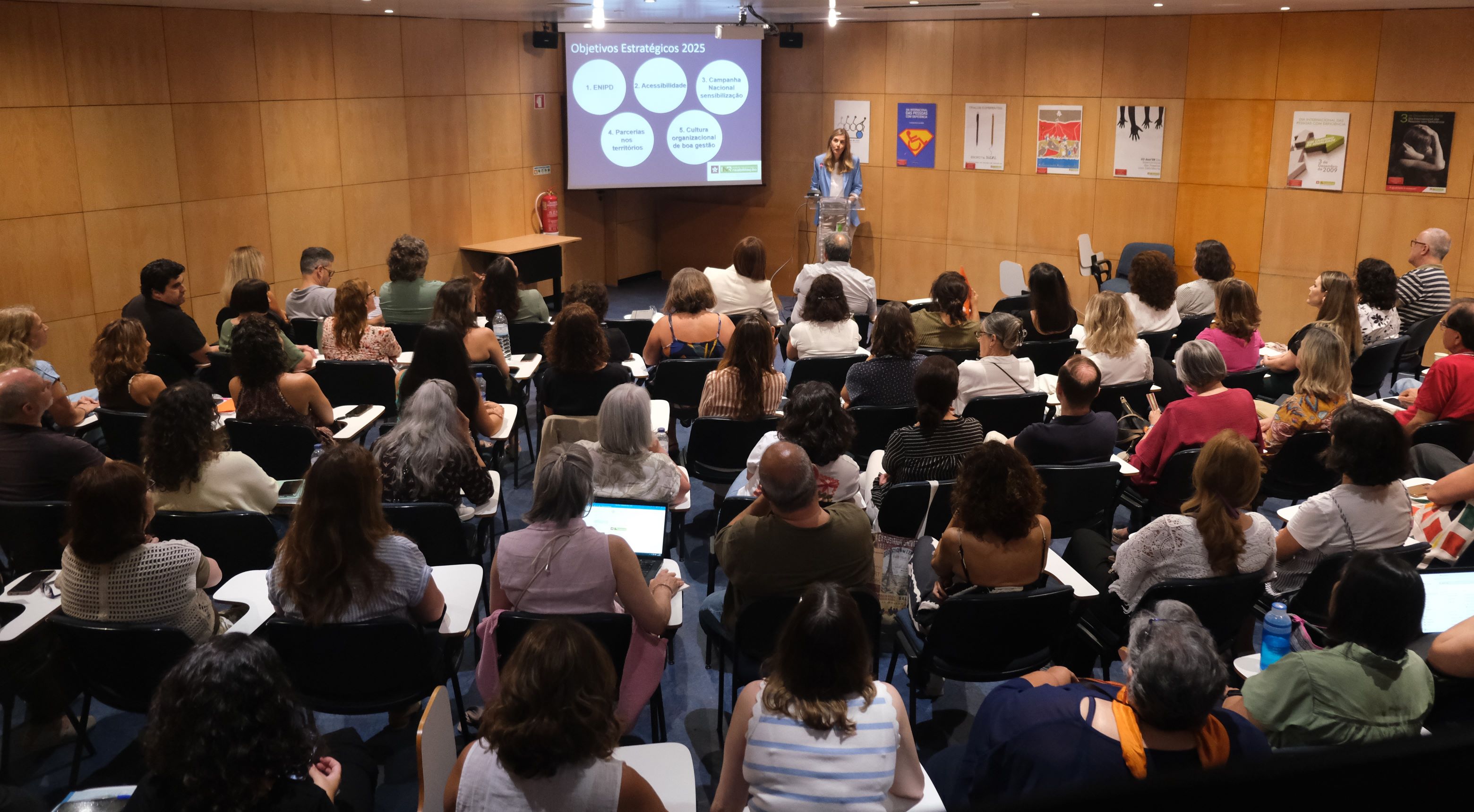 Auditório cheio com várias pessoas sentadas, voltadas para a oradora que fala no púlpito. Do lado esquerdo está uma tela a projetar um slide com o título 'Objetivos Estratégicos 2025' e vários pontos em lista. As paredes da sala são de madeira e têm vários cartazes afixados.