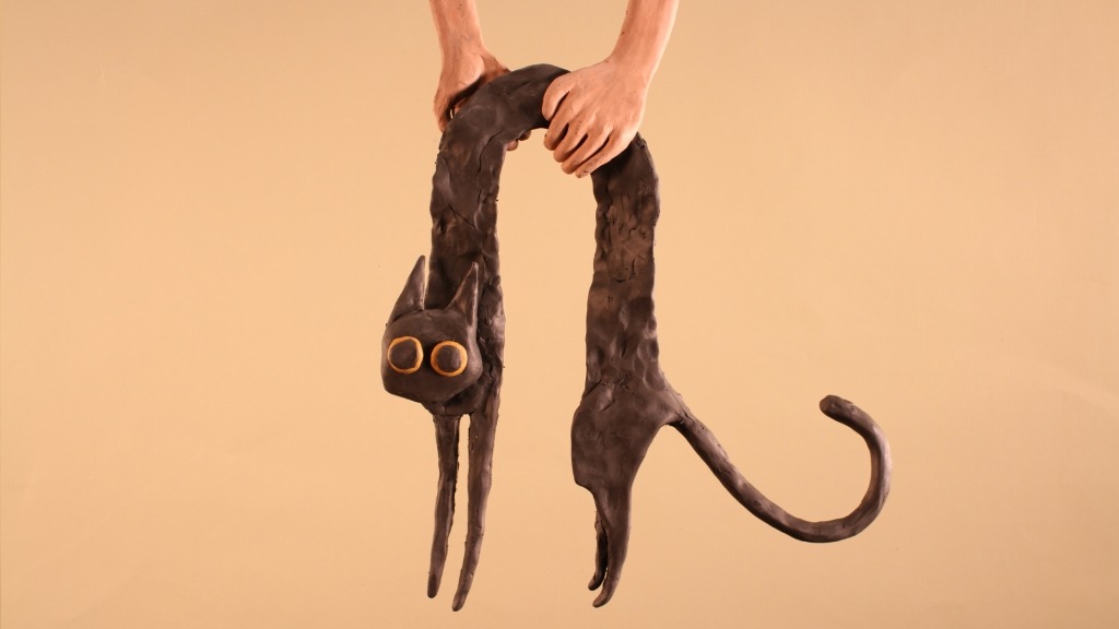 Duas mãos seguram uma escultura de barro preta em forma de gato, com pernas e cauda exageradamente alongadas. O gato tem olhos grandes e redondos de cor amarela e orelhas pontiagudas. O fundo é de cor bege uniforme
