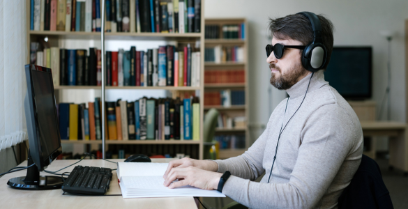 Homem sentado à secretária em frente a um monitor de computador, de óculos escuros e auscultadores. Ele está a ler um livro em Braille. Ao fundo, veem-se estantes com livros. 