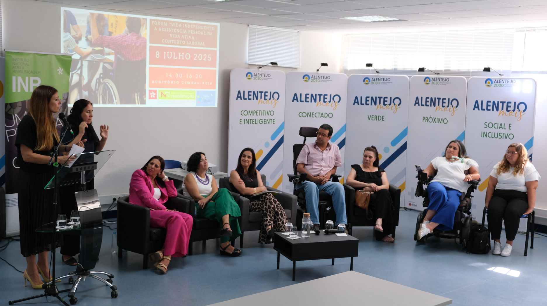 Seven people, participants in the forum, are seated. Two of them are in a wheelchair. On the left, standing, is a Portuguese Sign Language interpreter and speaking in the pulpit is Sónia Esperto, President of the INR. In the background, there are screens with the texts: 