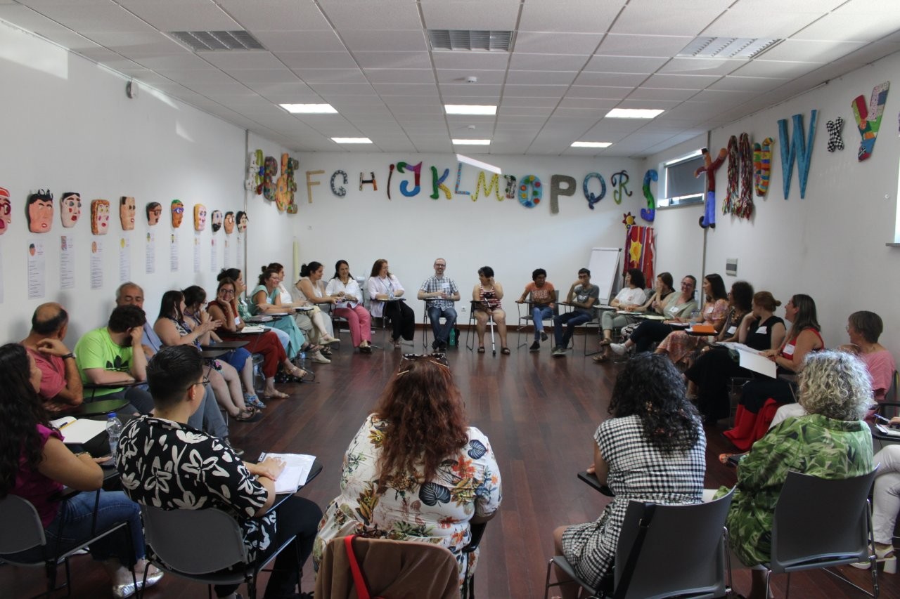  Grupo de pessoas sentadas em círculo numa sala decorada com letras do alfabeto e desenhos de rostos nas paredes. Algumas pessoas seguram papéis ou cadernos. Group of people sitting in a circle in a room decorated with letters of the alphabet and drawings of faces on the walls. Some people hold papers or notebooks. 