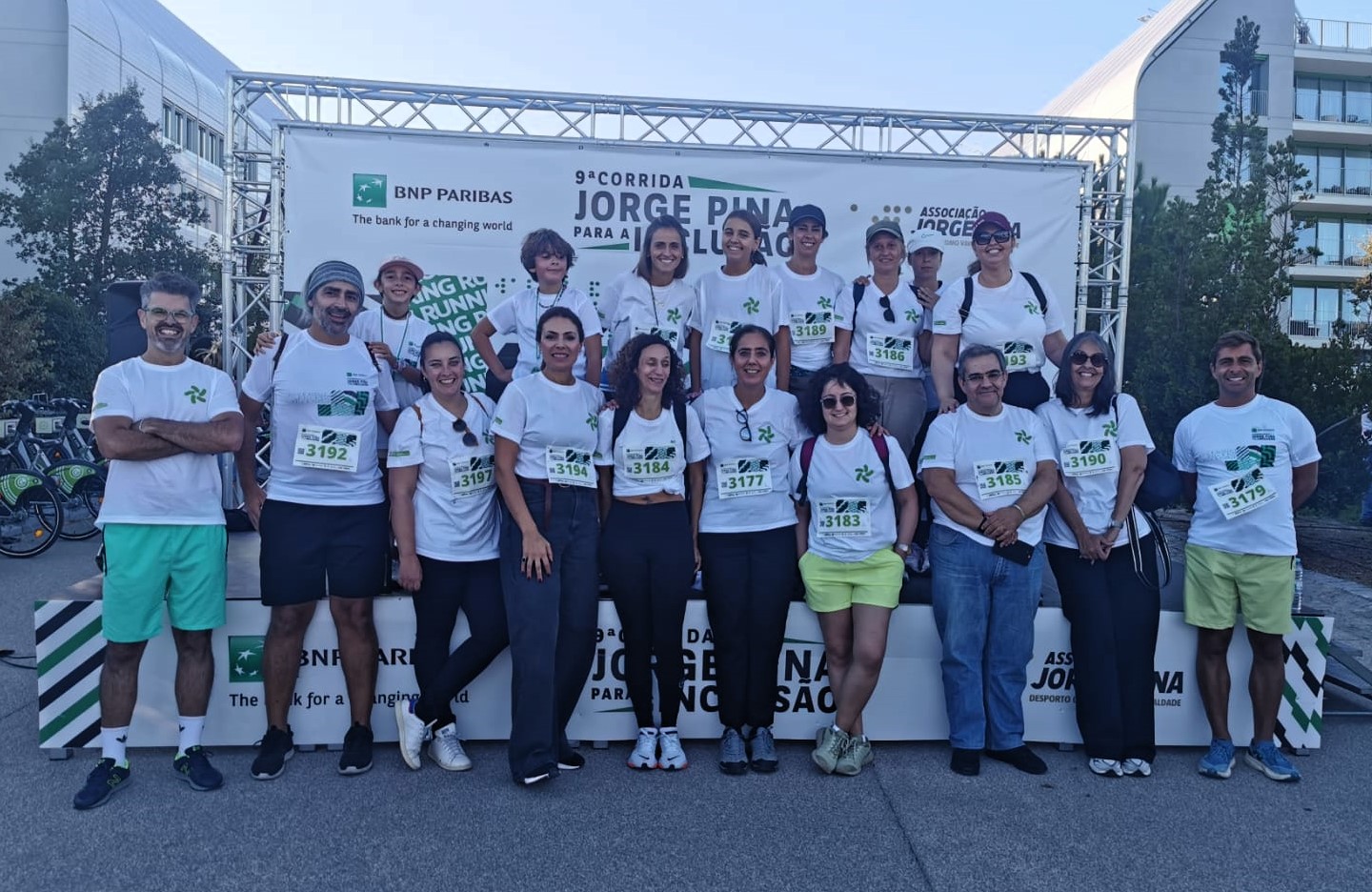  Um grupo de pessoas está posicionado em frente a um painel de evento ao ar livre. O painel exibe logótipos e textos, incluindo “BNP Paribas”, “9ª Corrida Jorge Pina para a Inclusão” e “Associação Jorge Pina”. As pessoas usam camisolas brancas com números de dorsal visíveis, indicando participação numa corrida. Algumas estão em pé sobre uma plataforma, enquanto outras estão no chão, formando duas filas. Ao fundo, vêem-se edifícios modernos, árvores e bicicletas estacionadas. A group of people is positioned in front of an outdoor event panel. The panel displays logos and texts, including “BNP Paribas”, “9th Jorge Pina Race for Inclusion” and “Jorge Pina Association”. People wear white jerseys with visible dorsal numbers, indicating participation in a race. Some are standing on a platform, while others are on the ground, forming two rows. In the background, you can see modern buildings, trees and parked bicycles. 