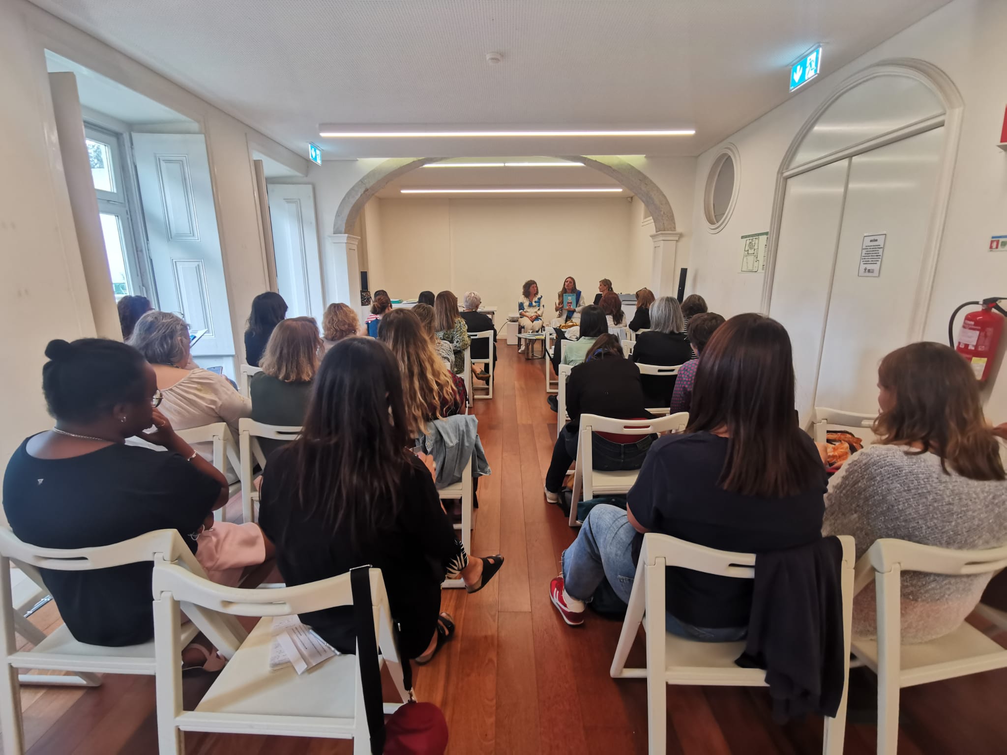 Rectangular room with light walls and wooden floor. The chairs are arranged in rows facing a table in front of the room. On the table are three people sitting, one of them holding a book in his hand. The audience is sitting in white chairs. On the left, there are large windows with natural light coming in.