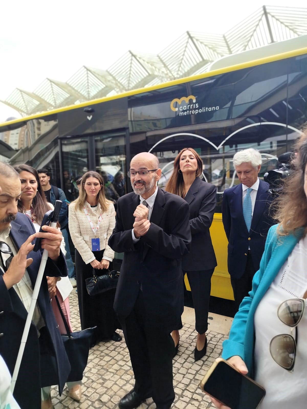  Grupo de pessoas em pé numa área exterior pavimentada, junto a um autocarro amarelo com a inscrição “Carris Metropolitana” na lateral. O autocarro está parado e ocupa grande parte do fundo da imagem. Por trás do veículo, vêem-se estruturas metálicas com cobertura translúcida, típicas de uma estação ou terminal. Algumas pessoas seguram telemóveis e outras estão com as mãos juntas à frente do corpo. No centro da imagem, uma pessoa fala para o grupo. 