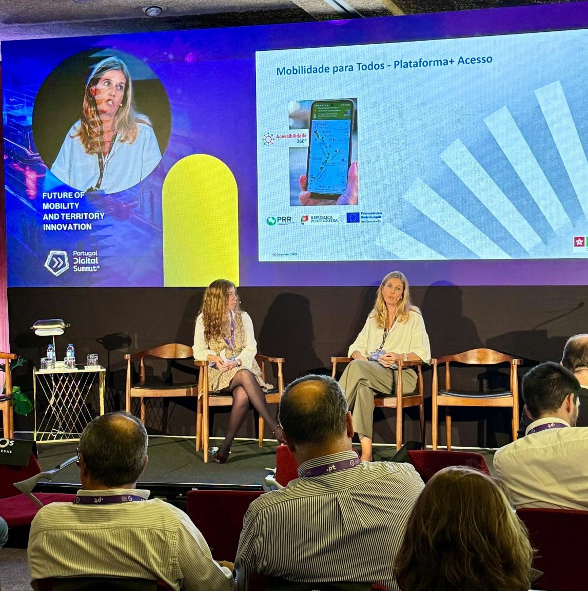 You see two people sitting there, one of them talking. Behind this is a big projection: On the left side, on a blue background, is a circle, with a photograph of one of the people who is sitting, and the rest is a photograph of a mobile phone. Above this image is the text “Mobility for All – Platform + Access”. It is also visible public sitting, watching.