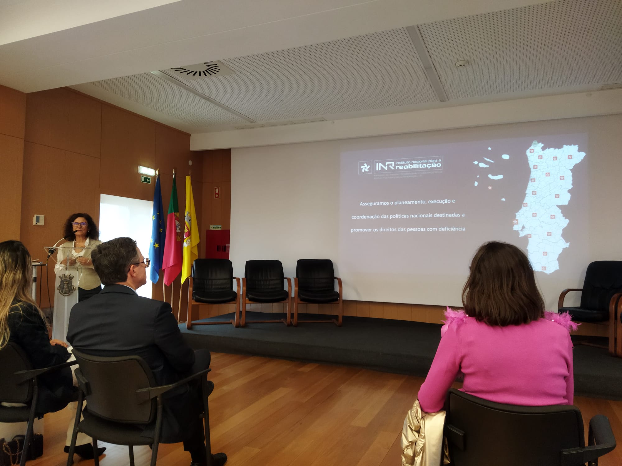  Sala de conferências com várias pessoas sentadas a assistir a uma apresentação. À esquerda, uma pessoa está de pé junto a um púlpito com o logótipo da instituição. Atrás do púlpito estão três bandeiras: União Europeia, Portugal e do Município de Mafra. No centro da imagem, vêem-se quatro cadeiras vazias alinhadas. No ecrã projetado aparece o logótipo do INR e o seguinte texto: 