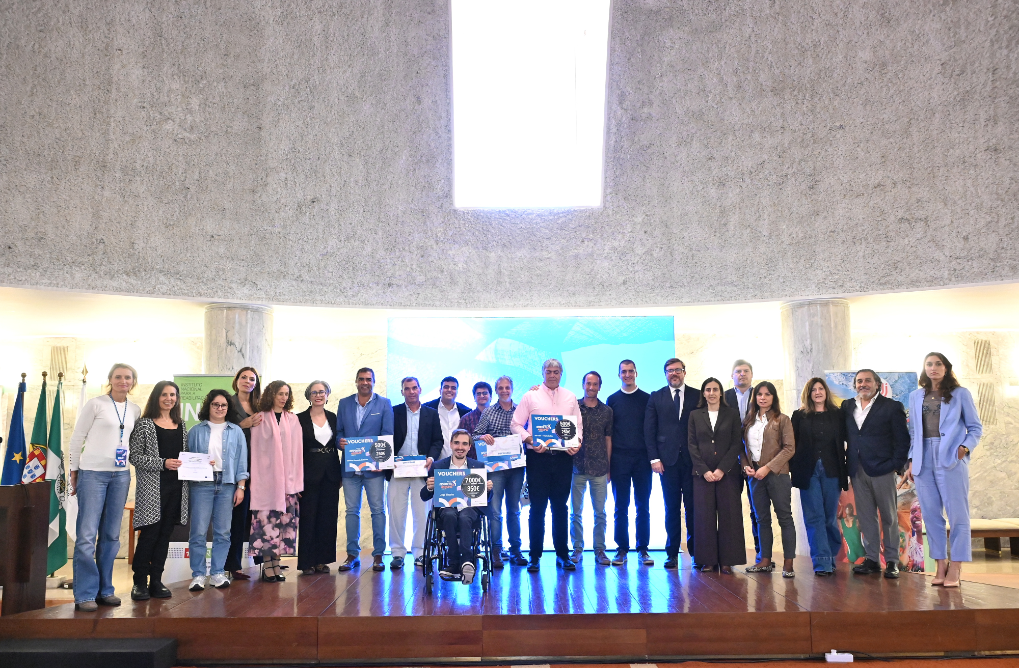 Grupo de pessoas posicionado em palco, formando uma linha. Ao centro, algumas seguram certificados e placas com texto visível. Uma pessoa está sentada numa cadeira de rodas à frente do grupo, também com uma placa. À esquerda, estão visíveis bandeiras, incluindo a de Portugal. Ao fundo, encontra-se uma parede curva de cor clara e um ecrã azul parcialmente visível.