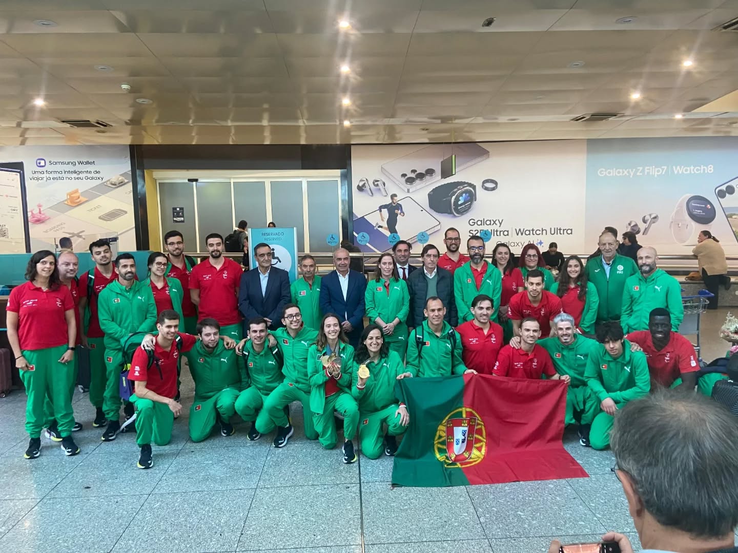  Grupo de pessoas posicionado em ambiente interior, junto a uma parede com publicidade a dispositivos eletrónicos. A maioria veste fatos desportivos verdes e vermelhos, e algumas pessoas usam roupa formal. Na parte frontal, duas pessoas seguram uma bandeira de Portugal. O grupo está organizado em várias filas, com algumas pessoas agachadas e outras de pé. Group of people positioned in an indoor environment, next to a wall with advertising for electronic devices. Most wear green and red sports suits, and some people wear formal clothing. On the front, two people hold a Portuguese flag. The group is organized in several rows, with some people squatting and others standing. 