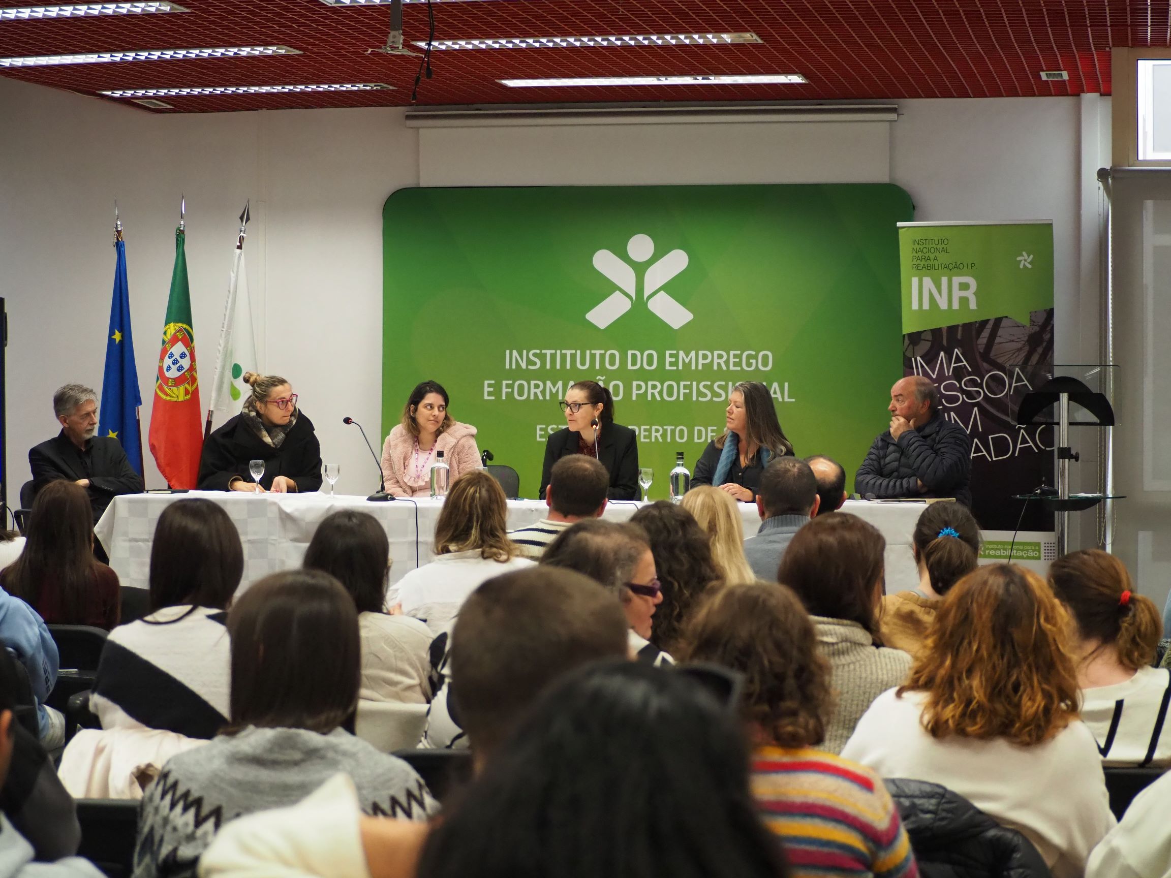 Auditório com várias pessoas sentadas a assistir a uma mesa-redonda composta por seis intervenientes. A mesa está coberta com toalha branca e equipada com microfones e copos de água. Atrás dos oradores, há um painel verde com o logótipo e a inscrição “Instituto do Emprego e Formação Profissional”. À esquerda, estão hasteadas as bandeiras da União Europeia, de Portugal e do IEFP. À direita, existe um rollup do INR.