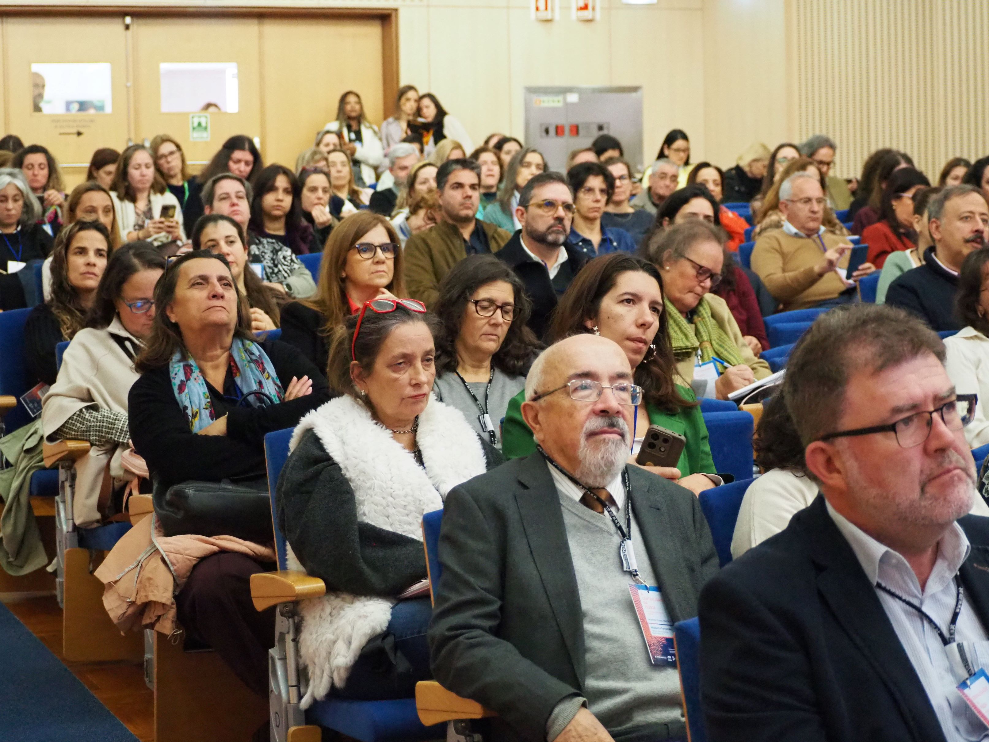 Auditório com várias filas de cadeiras azuis ocupadas por pessoas sentadas. Algumas seguram telemóveis ou blocos de notas. No fundo, há uma porta dupla de madeira clara e, perto dela, algumas pessoas estão de pé. As paredes são revestidas com painéis claros.