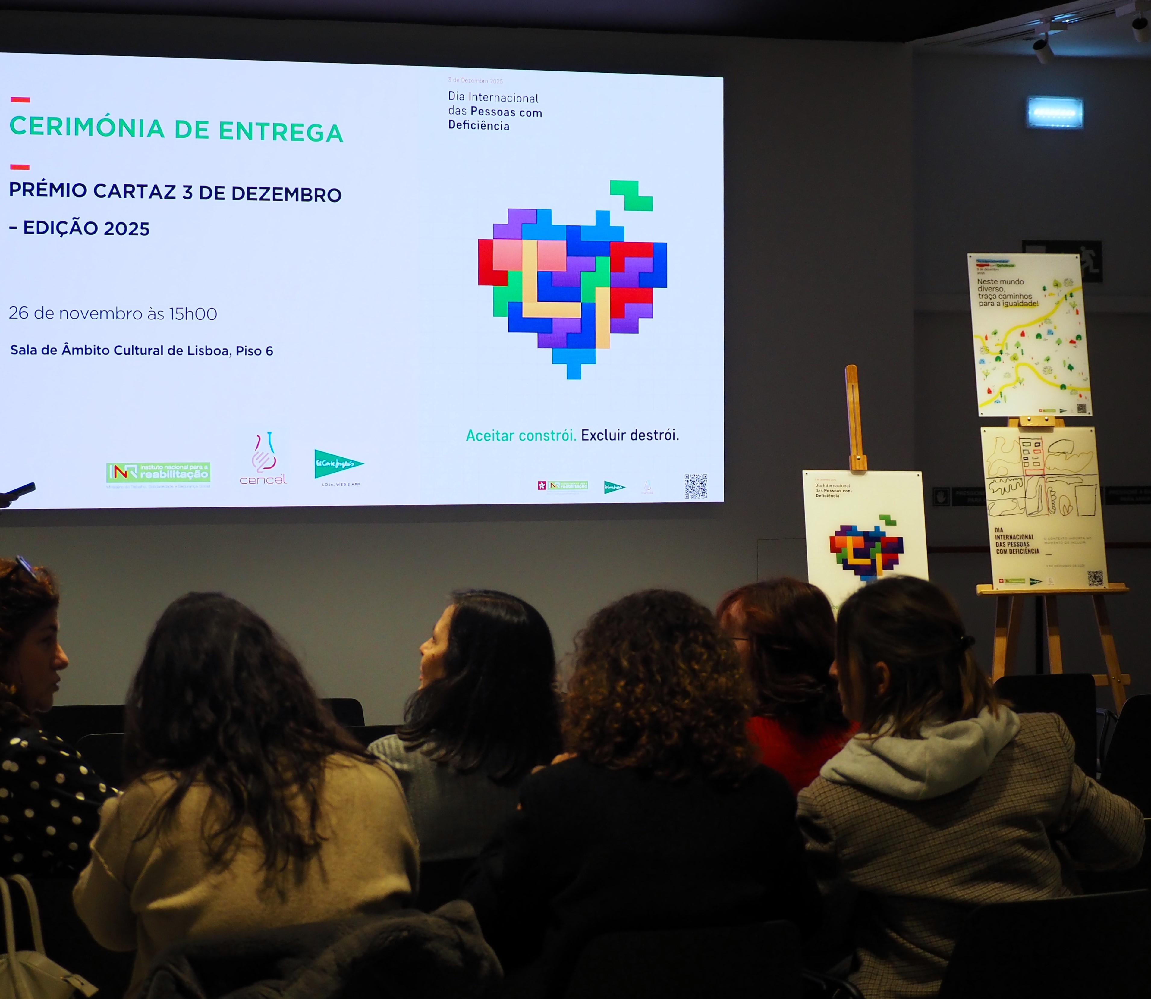 Fotografia da cerimónia de entrega do Prémio Cartaz 3 de Dezembro – Edição 2025, realizada numa sala com projeção em ecrã. No ecrã lê-se: “CERIMÓNIA DE ENTREGA Prémio Cartaz 3 de Dezembro – Edição 2025 26 de novembro às 15h00 Sala de Âmbito Cultural de Lisboa, Piso 6”. Ao centro do ecrã está um coração formado por blocos coloridos estilo Tetris. Na parte inferior, a frase: “Aceitar constrói. Excluir destrói.”. À direita, sobre cavaletes, estão os três cartazes premiados. Em primeiro plano, várias pessoas sentadas assistem à apresentação.