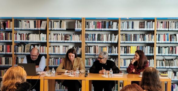  Fotografia do evento. Vê-se quatro pessoas sentadas numa mesa, atrás delas livros em estantes, e na sua frente outras pessoas sentadas. Photo of the event. You see four people sitting at a table, behind them books on shelves, and in front of them other people sitting. 