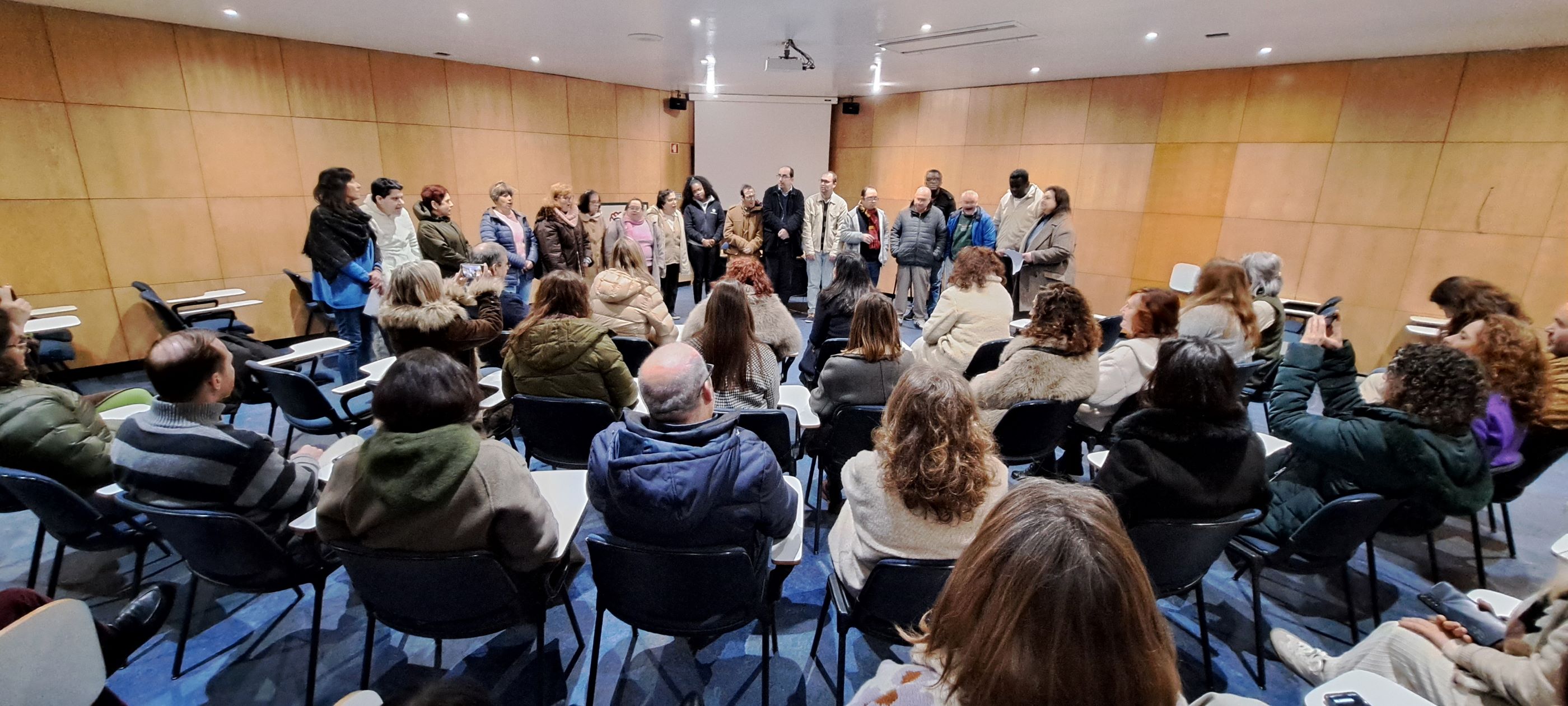  An auditorium is visible, with an audience present. You can see a group of 18 people standing in the background, singing. É visível um auditório, com público presente. Ao vê-se um fundo grupo de 18 pessoas em pé, a cantar. 