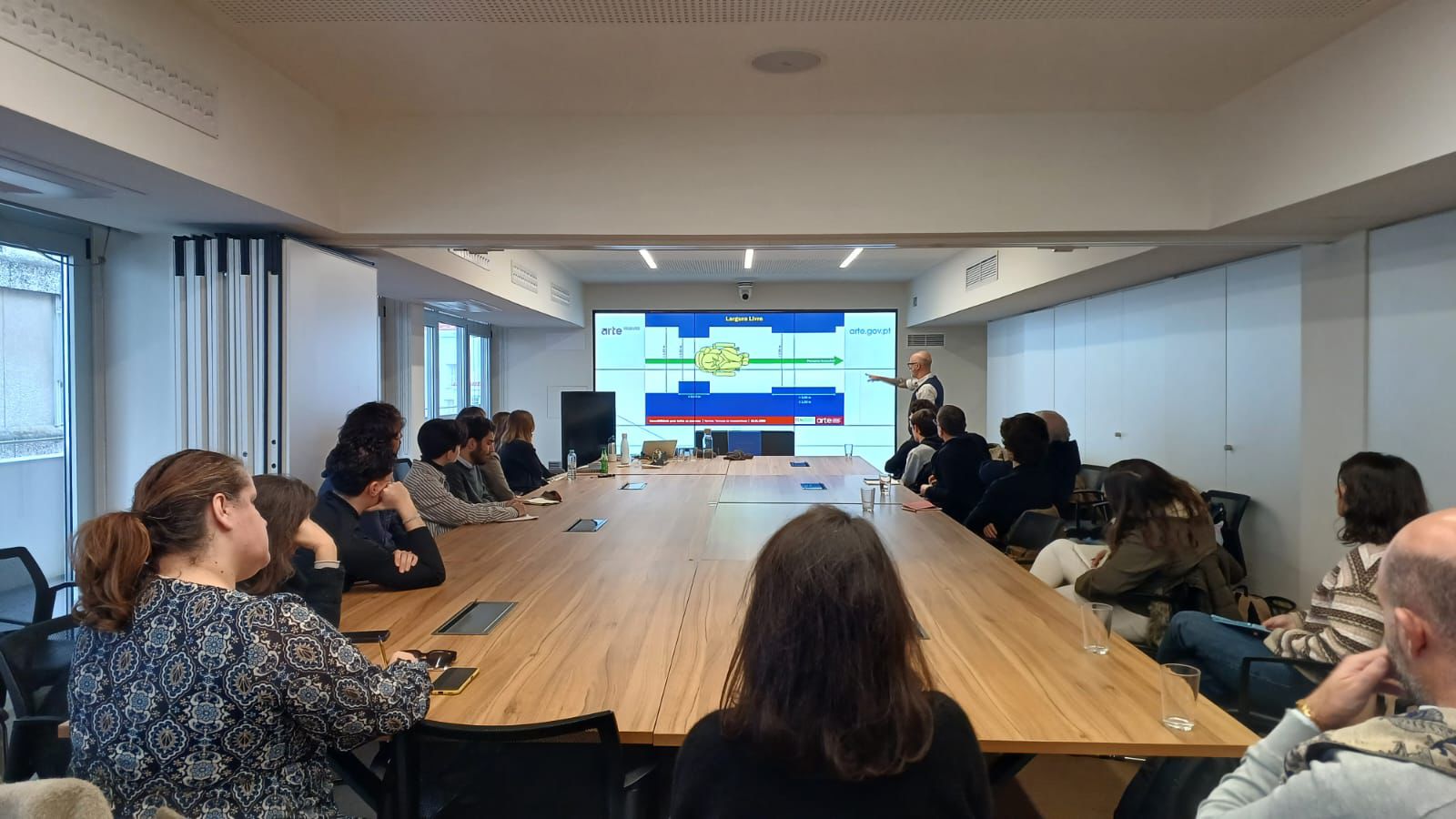  Uma sala de reuniões com uma mesa comprida de madeira, em torno da qual várias pessoas estão sentadas a assistir a uma apresentação. Ao fundo, um apresentador em pé aponta para um ecrã grande que mostra um slide com gráficos e blocos coloridos. A sala tem paredes claras, iluminação de teto e janelas laterais. A meeting room with a long wooden table, around which several people are seated to watch a presentation. In the background, a standing presenter points to a large screen that shows a slide with colorful graphics and blocks. The room has light walls, ceiling lighting and side windows. 