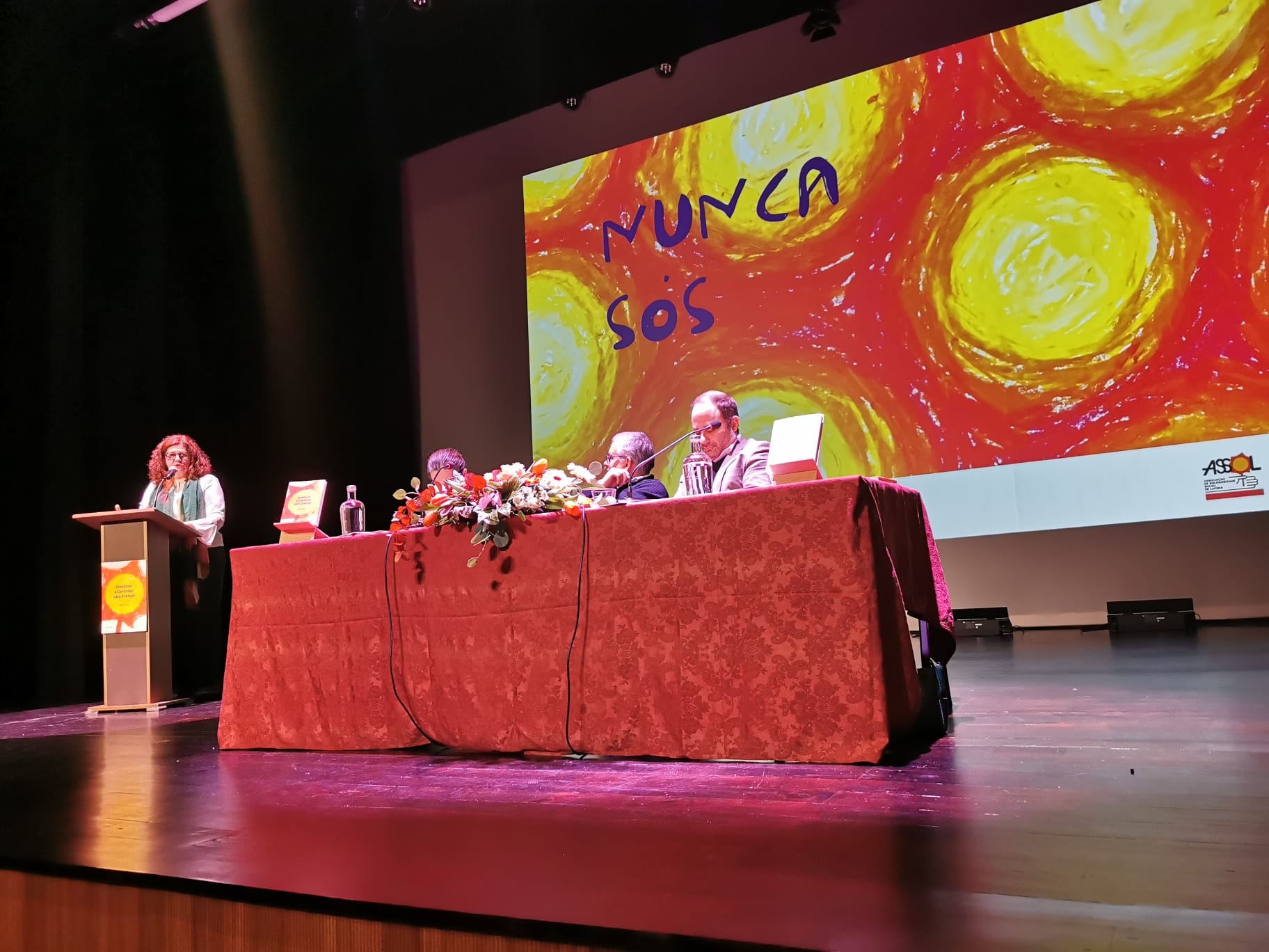 Palco de um auditório com uma mesa comprida coberta por toalha vermelha, onde estão sentadas várias pessoas com microfones, garrafas de água e arranjos florais. À esquerda, uma pessoa está em pé a falar num púlpito. Ao fundo, um ecrã projeta uma imagem abstrata em tons de amarelo e vermelho com as palavras “NUNCA SÓS” escrita à mão. O espaço está iluminado por focos de luz e apresenta um ambiente de conferência ou sessão pública.