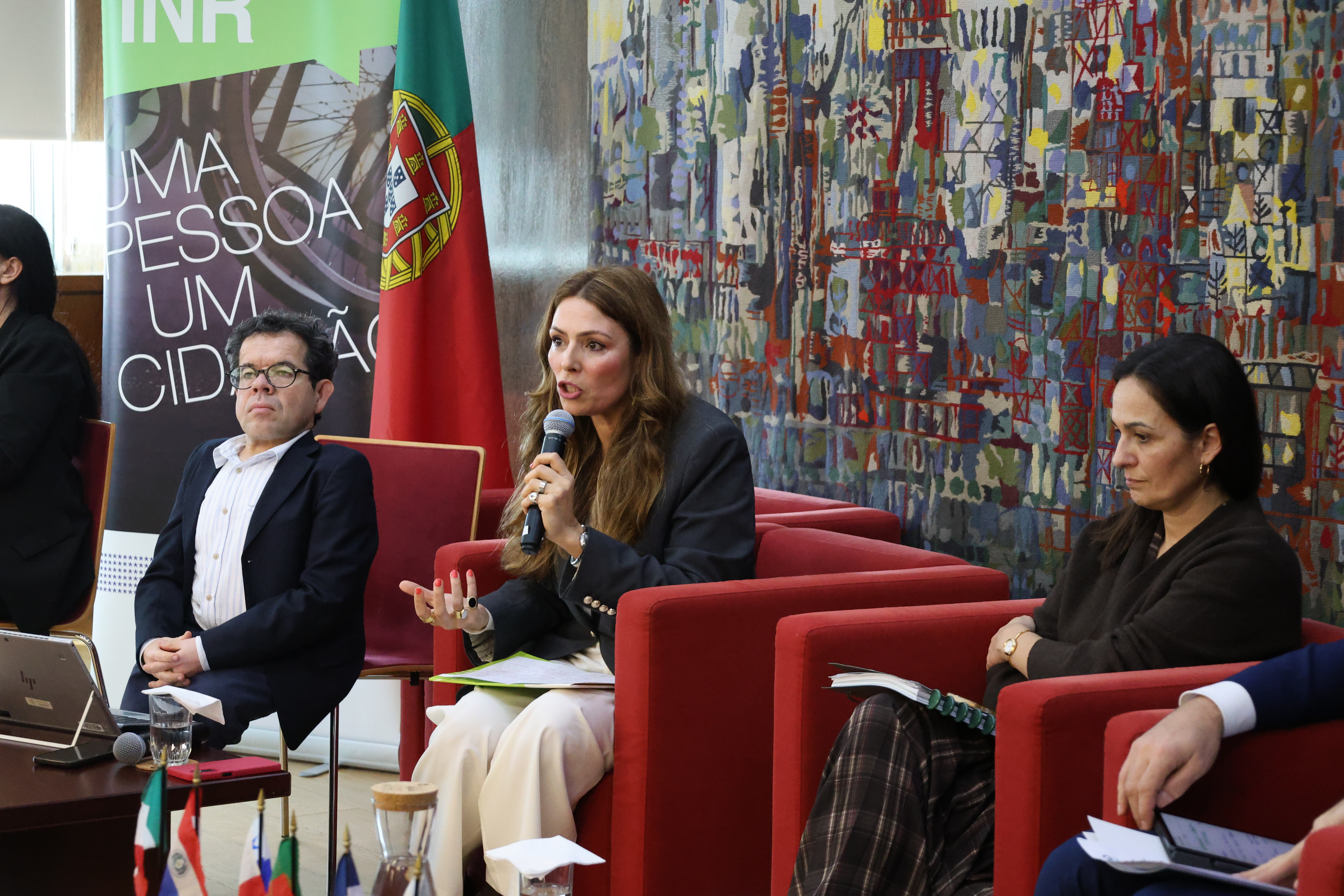  Esta imagem refere se ao momento Sónia Esperto, Presidente do Instituto Nacional para a Reabilitação (INR)., faz a sua intervenção no Seminário sobre a Promoção da Autonomia e Vida Independente, no âmbito do Programa Ibero Americano de Deficiência, promovido pelo INR, a 24 de fevereiro de 2026, no Salão Nobre do Ministério do Trabalho, Solidariedade e Segurança Social, em Lisboa. Na imagem vê-se várias pessoas sentadas em cadeirões vermelhos, dispostas em linha, uma mesa redonda. Sobre a mesa existe documentação e pequenos conjuntos de bandeiras. Atrás do grupo, vê se uma tapeçaria multicolorida de grande dimensão e, à esquerda, um cartaz institucional com o texto “INR – UMA PESSOA, UM CIDADÃO”, bem como uma bandeira de Portugal colocada verticalmente. This image refers to the moment Sónia Esperto, President of the National Institute for Rehabilitation (INR)., makes his speech at the Seminar on the Promotion of Autonomy and Independent Living, under the Ibero-American Disability Program, promoted by the INR, on February 24, 2026, at the Noble Hall of the Ministry of Labor, Solidarity and Social Security, in Lisbon. The image shows several people sitting in red armchairs, arranged in line, a round table. On the table there is documentation and small sets of flags. Behind the group is a large multi-coloured tapestry and, on the left, an institutional poster with the text ‘INR – ONE PERSON, ONE CITIZEN’, as well as a vertically placed Portuguese flag. 