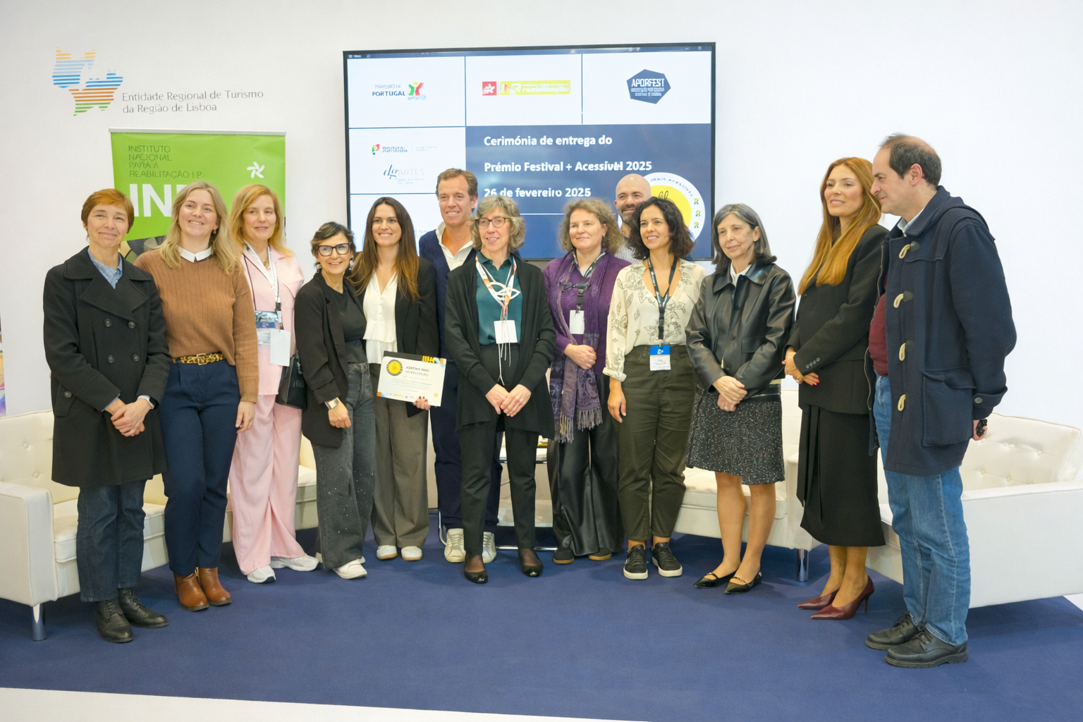 Um grupo de 12 pessoas está alinhado numa sala durante uma cerimónia de entrega do Prémio Festival+ Acessível 2025. Ao fundo, um grande ecrã exibe informações sobre o evento, acompanhado por vários logótipos institucionais. Uma pessoa segura um certificado. O espaço inclui sofás brancos nas laterais e um chão azul‑escuro.