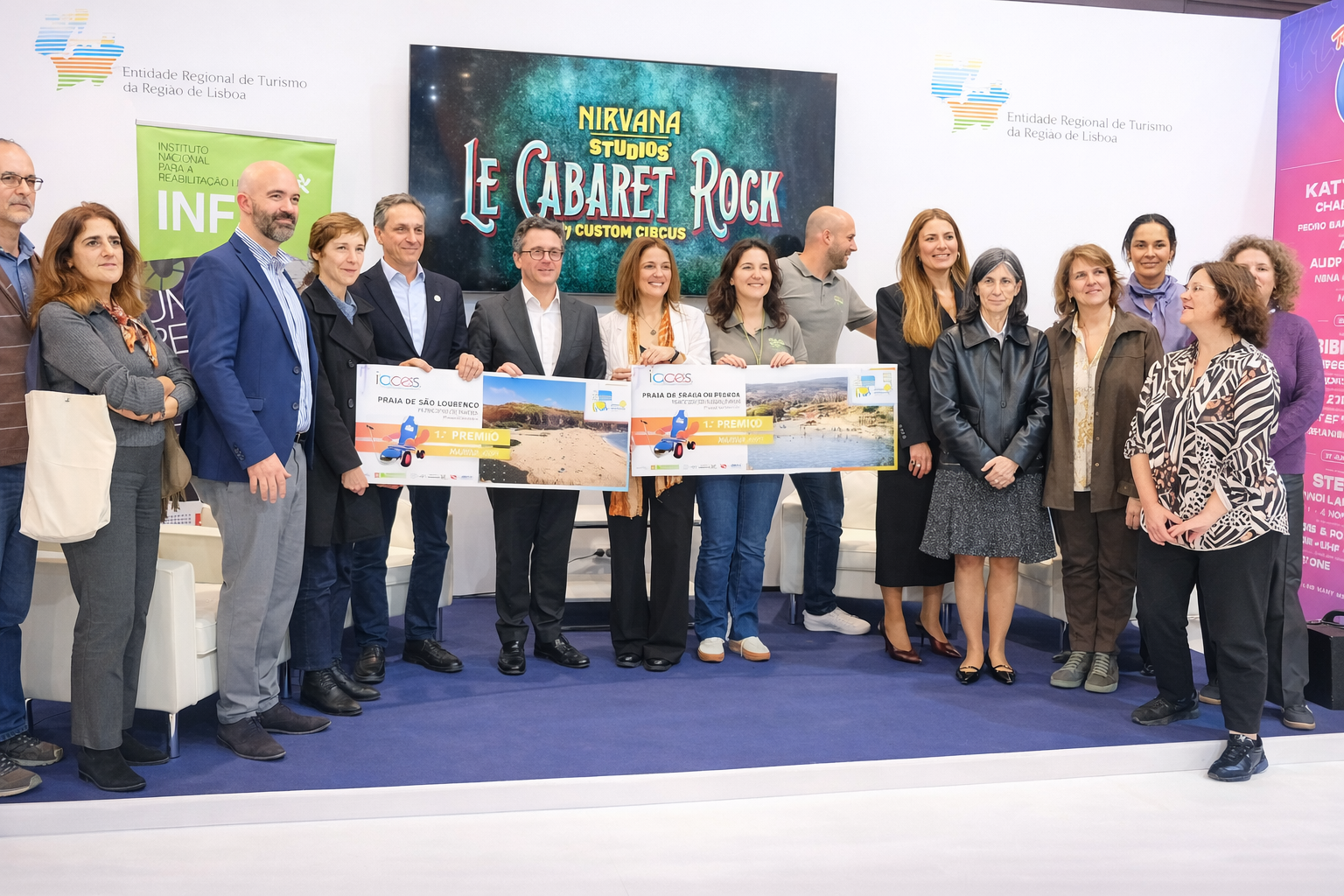 Um grupo de pessoas está reunido num espaço de exposição durante uma cerimónia relacionada com o Prémio ‘Praia + Acessível’ 2025. No centro, algumas pessoas seguram duas placas com as imagens das praias e textos alusivos ao prémio. À esquerda e à direita estão painéis verticais com logótipos e informação institucional. O chão é azul escuro e existem sofás brancos ao fundo. A iluminação é clara, típica de um evento em recinto fechado.