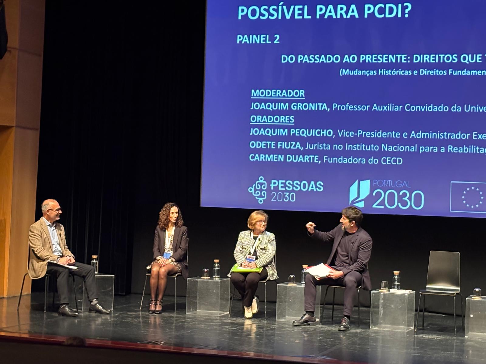  Palco de um auditório com quatro pessoas sentadas em cadeiras, dispostas em linha, durante um painel de debate. Atrás, um ecrã projeta a identificação do “Painel 2”, dedicado aos direitos das pessoas com deficiência, com indicação do moderador e dos oradores, bem como os logótipos “Pessoas 2030”, “Portugal 2030” e da União Europeia. Um dos participantes está a intervir, enquanto os restantes escutam. Stage of an auditorium with four people sitting in chairs, arranged online, during a panel discussion. Behind it, a screen projects the identification of the ‘Panel 2’, dedicated to the rights of persons with disabilities, with an indication of the moderator and speakers, as well as the ‘People 2030’, ‘Portugal 2030’ and European Union logos. One of the participants is intervening, while the others are listening. 