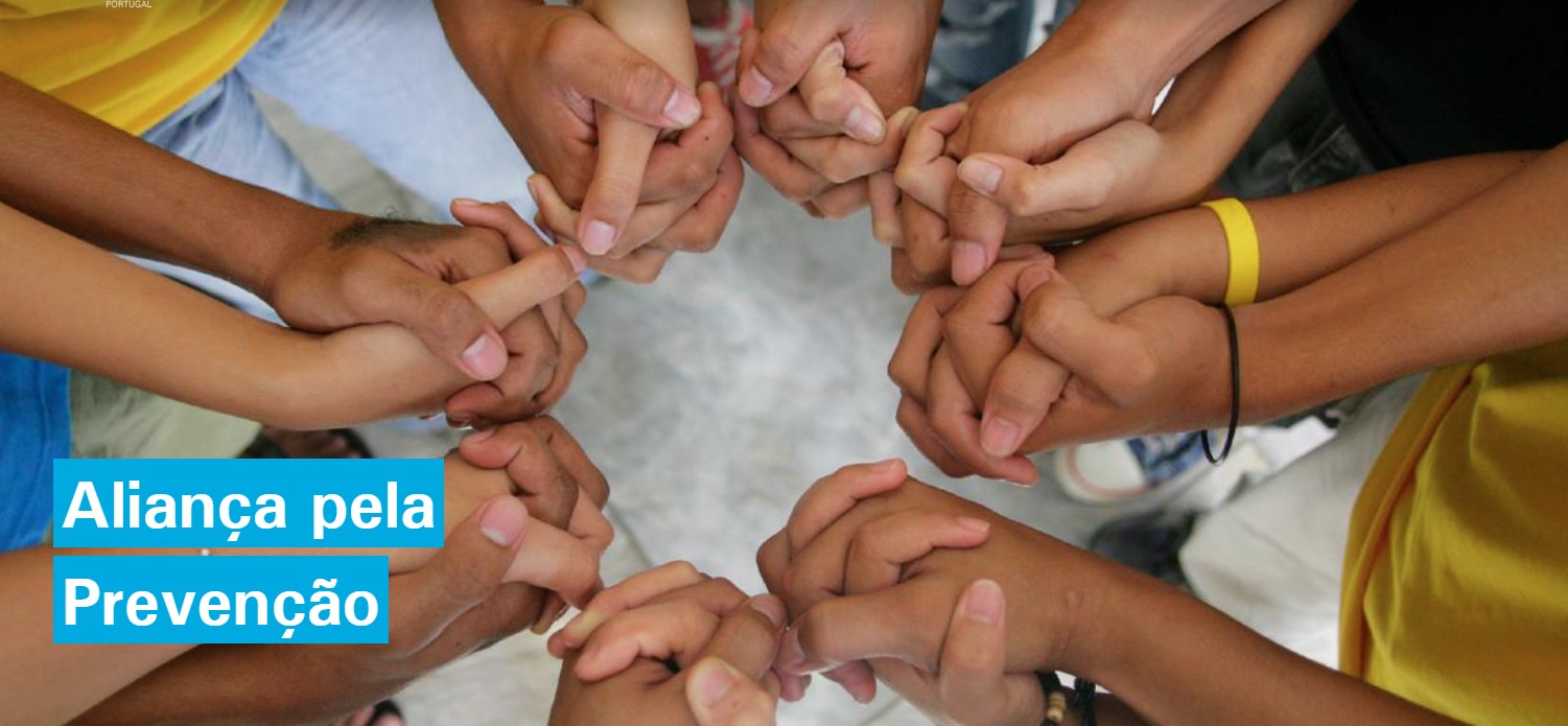 Um grupo de várias pessoas forma um círculo, unindo ambas as mãos entrelaçadas com as mãos das pessoas ao lado. Apenas os antebraços e mãos estão visíveis. As mãos estão posicionadas no centro da imagem, criando um padrão circular. O fundo é neutro e indistinto. No lado esquerdo, sobre um retângulo azul, aparece o texto: “Aliança pela Prevenção”.