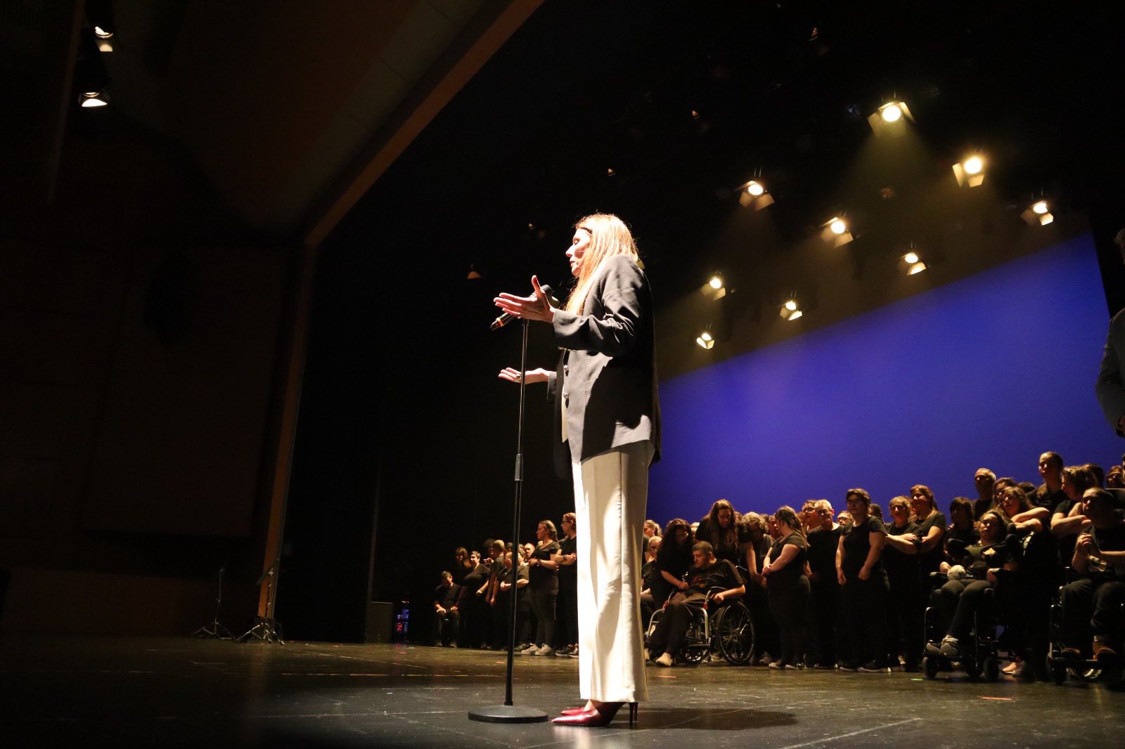  Palco de teatro com iluminação superior direcionada. Em primeiro plano, uma pessoa está de pé junto a um microfone, com um braço levantado e o outro parcialmente aberto, voltada para o público. No fundo do palco, um grupo numeroso de pessoas está alinhado, algumas de pé e pelo menos uma em cadeira de rodas. O cenário é marcado por um fundo liso de cor azul e pela presença de focos de luz no teto. Theater stage with directed superior lighting. In the foreground, one person stands next to a microphone, with one arm raised and the other partially open, facing the public. At the back of the stage, a large group of people are lined up, some standing up and at least one in a wheelchair. The scenery is marked by a smooth background of blue color and the presence of light spots on the ceiling. 