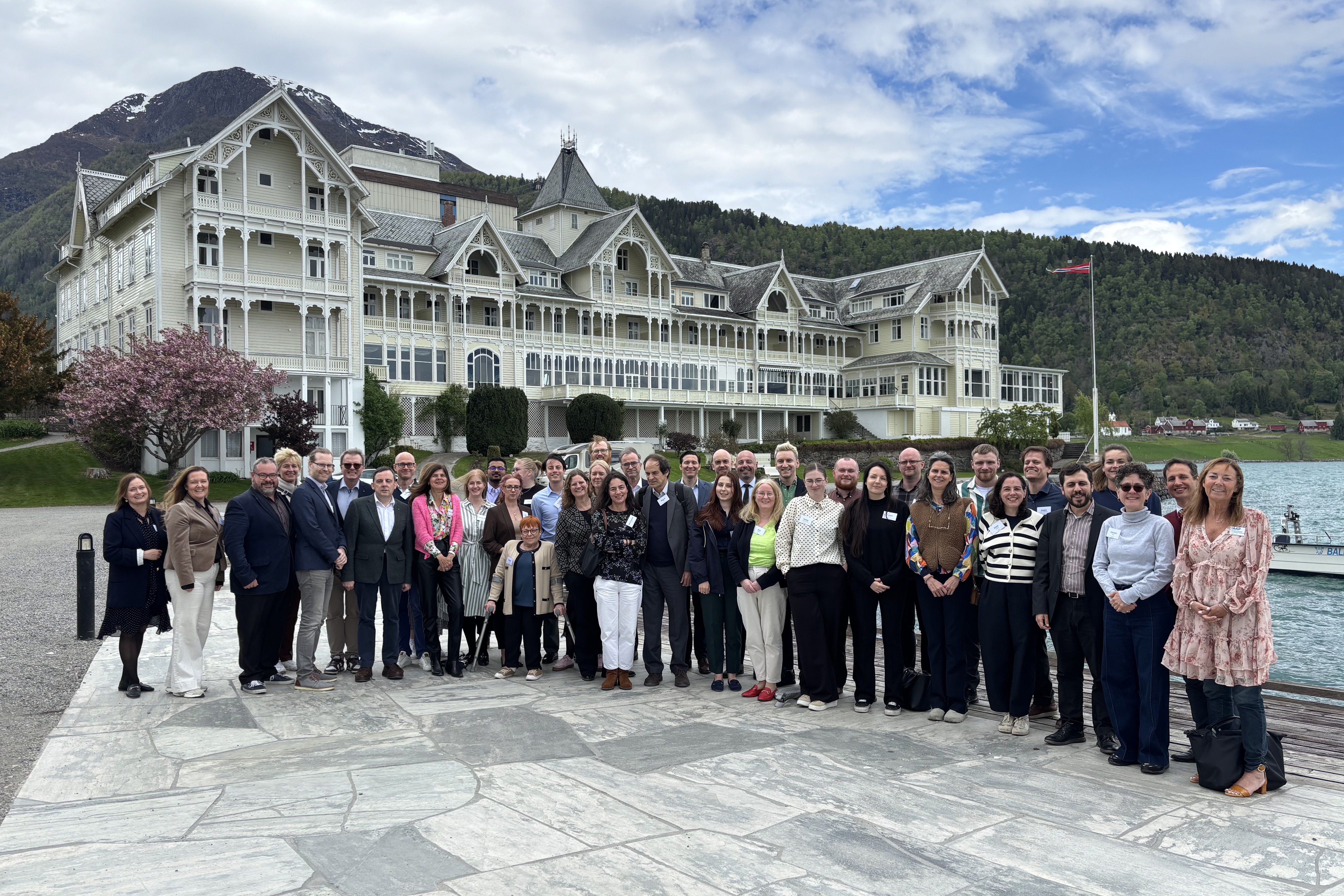  Fotografia de grupo ao ar livre com cerca de três dezenas de pessoas alinhadas e voltadas para a câmara, sobre um pavimento em pedra junto a um lago. Ao fundo encontra-se um edifício grande, de estilo histórico. Outdoor group photo with about three dozen people lined up and facing the camera, on a stone pavement next to a lake. In the background is a large, historical-style building. 