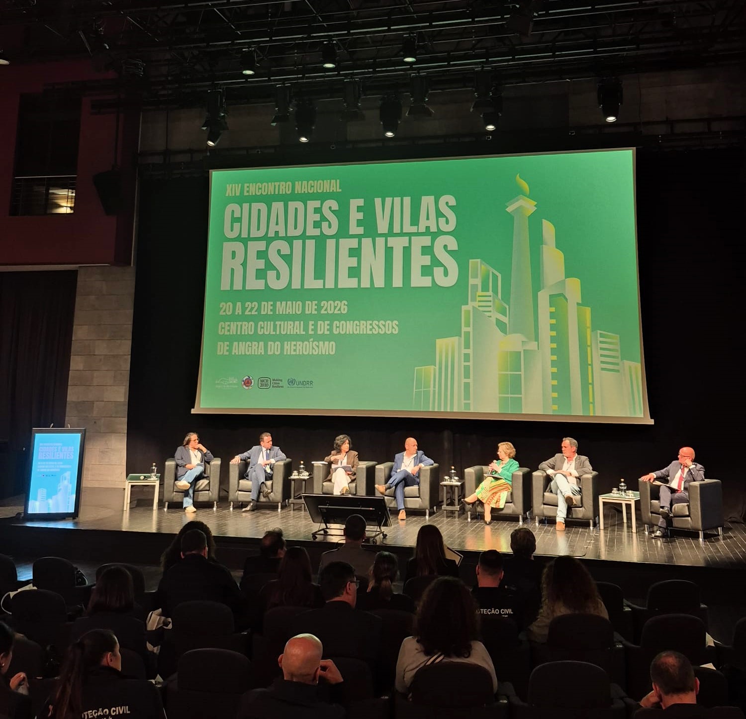 Fotografia de um auditório durante um evento. No palco, várias pessoas estão sentadas em cadeiras dispostas em linha, a participar num painel de debate. À frente do palco encontram-se cadeiras ocupadas pelo público. Ao fundo, um grande ecrã projeta um fundo verde com texto em branco que diz: “XIV Encontro Nacional – Cidades e Vilas Resilientes”. O ecrã inclui ainda a informação “20 a 22 de maio de 2026” e “Centro Cultural e de Congressos de Angra do Heroísmo”. À direita do texto, há uma ilustração estilizada de edifícios urbanos.