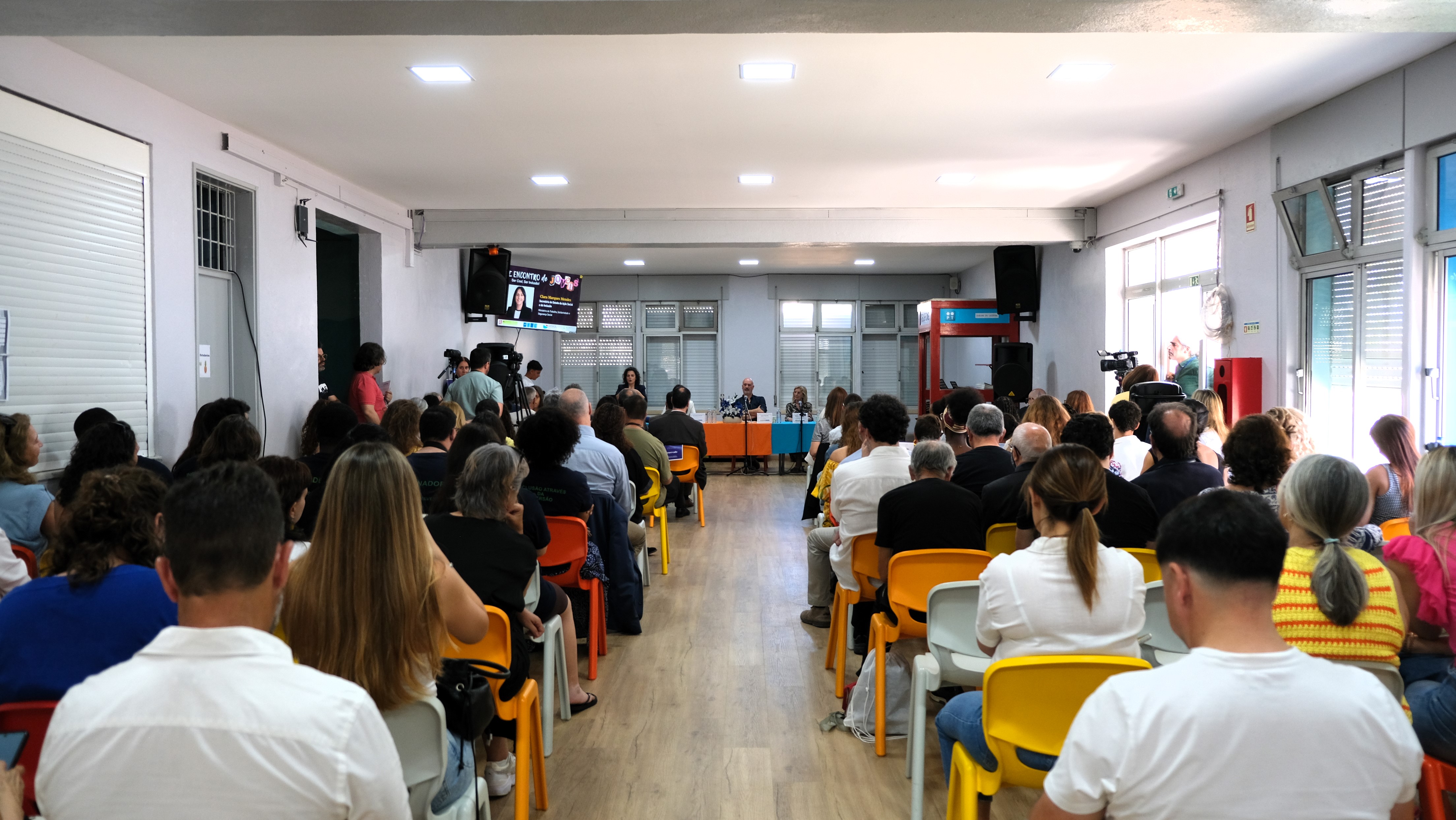 View of the room with several rows of chairs occupied by participants facing a table where a panel takes place. A screen is visible on the wall in the background.