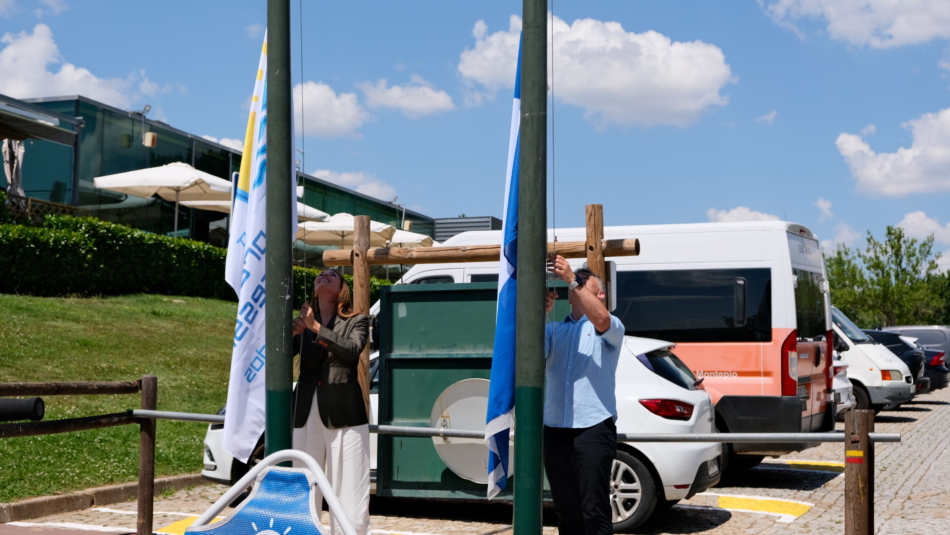  Duas pessoas junto a mastros puxam cordas para hastear bandeiras, num espaço exterior. Two people next to masts pull strings to fly flags in an outdoor space. 