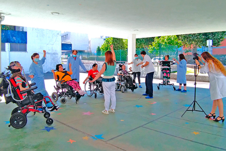 No exterior de um edifício, numa área coberta, de chão verde com estrelas coloridas, estão algumas Pessoas, alternadamente em pé e em cadeira de rodas. Ao centro está uma Mulher de costas, de braço no ar e, atrás desta, outra Mulher, inclinada sobre uma máquina fotográfica, em cima de um tripé.