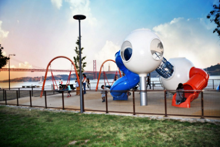 Parque infantil com crianças a brincar em baloiços e escorregas tubulares ligados por duas bolas.