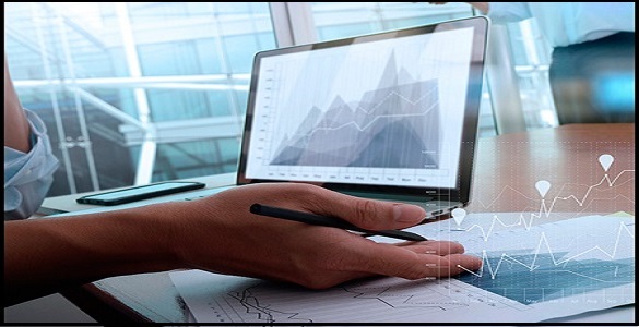 secretária sobre a qual se vê um portátil aberto, um telemóvel, vários papeis e uma mão que segura uma caneta