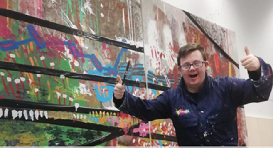 Um jovem com Síndrome de Down posa com os polegares levantados à frente de uma enorme tela abstrata e colorida
