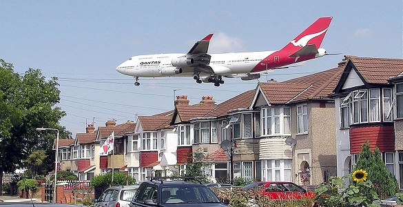 Rua com um conjunto de casas geminadas e carros estacionados e um avião que quase rasa os telhados.