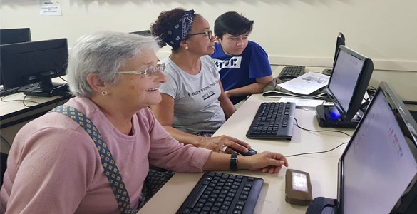 Uma mulher idosa, uma mulher de meia idade e um jovem sentados à frente de computadores.