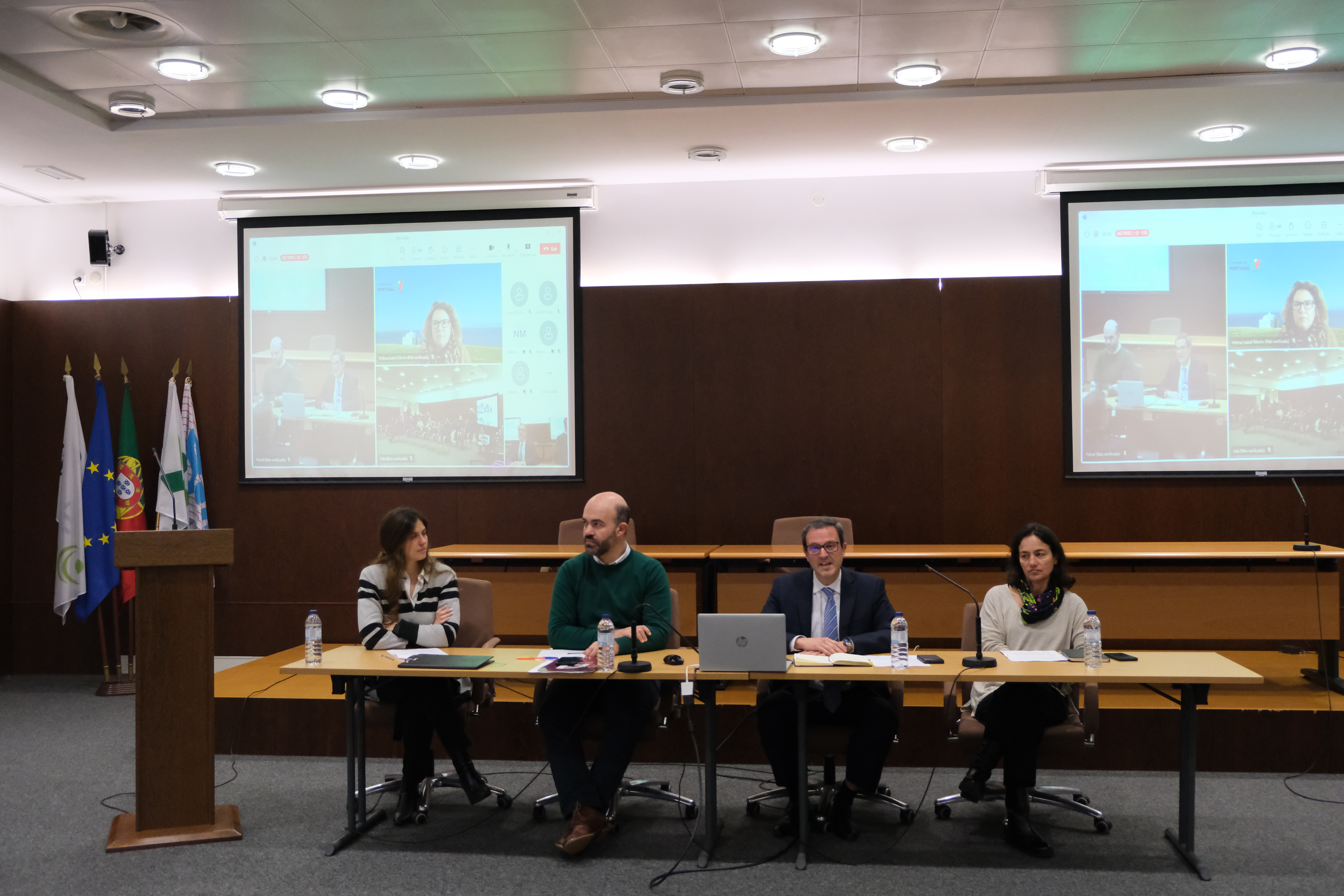 A imagem mostra uma mesa com quatro pessoas (Alexandra Andrade, Nelson Nunes, Rodrigo Ramos - Presidente e Ana Martins, pertencentes ao Instituto Nacional para a Reabilitação) numa sala de conferências. Por detrás das pessoas estão duas telas projetadas, com várias janelas mostrando participantes remotos.