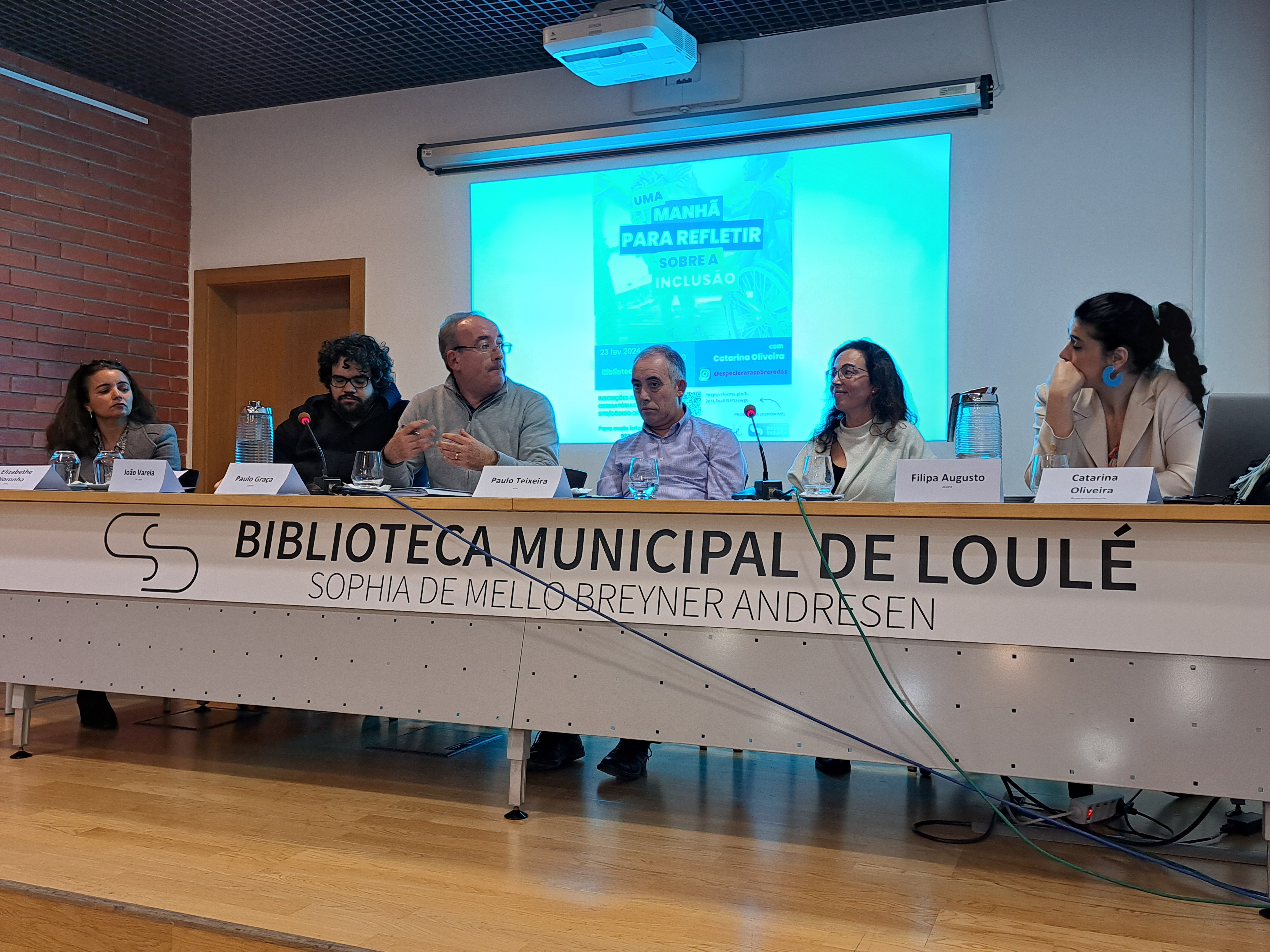 Legend: Discussion session of the event “A morning to reflect on Inclusion” held (from right to left) Catarina Oliveira, moderator; Filipa Augusto, ACAPO; Paulo Teixeira, APPC; Paulo Graça, EXIST; João Varela, CVI and Elisabethe Noronha, ASMAL at the Municipal Library of Loulé. Audio description: Projection on the wall on the theme. Table with six people, Paulo Graça is talking and gesturing to Catarina before the attention of the others.