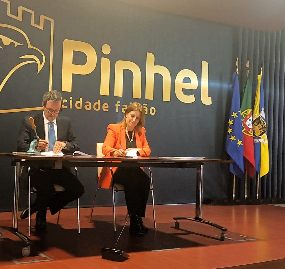 A foto mostra um palco com um pano de fundo azul escuro exibindo o texto “Pinhel Cidade Falcão”. Duas pessoas estão sentadas atrás de uma mesa longa a assinar o Protocolo de Cooperação. À direita do palco está a Senhora Vice-Presidente da Câmara Municipal de Pinhel e, à esquerda, o Senhor Presidente do Conselho Diretivo do INR. Há três bandeiras montadas em postes: a da União Europeia, a bandeira de Portugal e uma bandeira amarela e azul com o brasão da cidade.