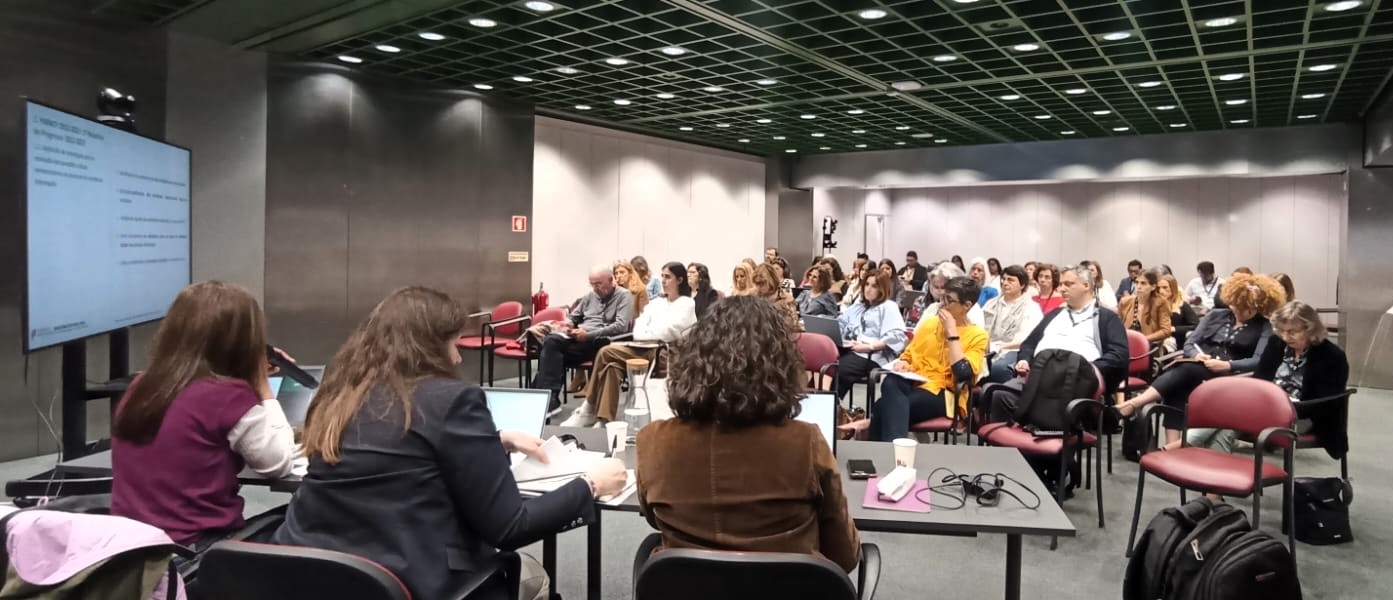 A imagem mostra uma sala de conferências com pessoas sentadas em filas, de frente para uma televisão que exibe uma apresentação e para uma mesa com 3 oradores, em que um deles está a falar através de um microfone.